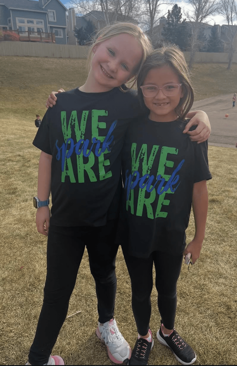Two young girls with glasses and long hair, smiling and hugging outdoors on grass, wearing matching black shirts with green and blue text.