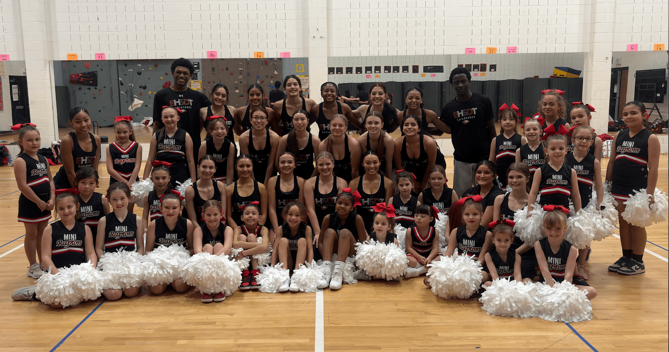 Group of cheerleaders and coaches posing indoors, some holding pom-poms, in front of a wall with lockers.