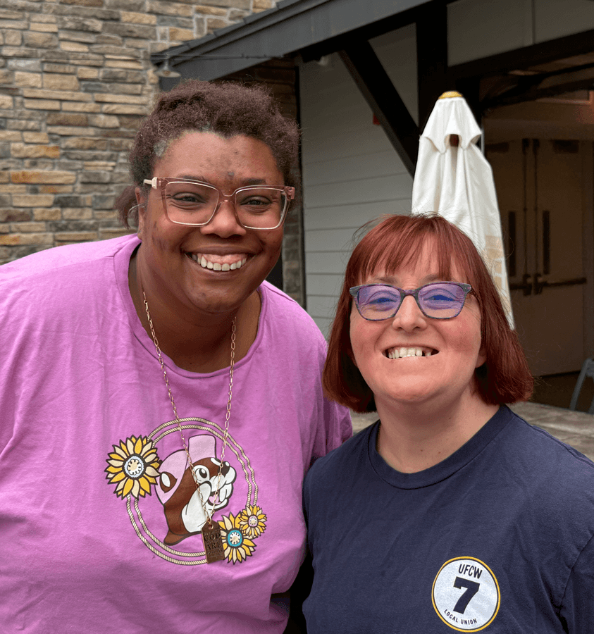 Two smiling women with glasses, one in a pink shirt and the other in a navy shirt, outdoors near a building.