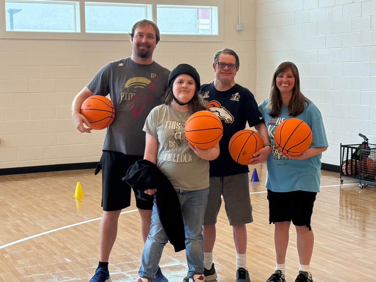 Four people standing in a gymnasium holding basketballs, with a basketball hoop and cones in the background.