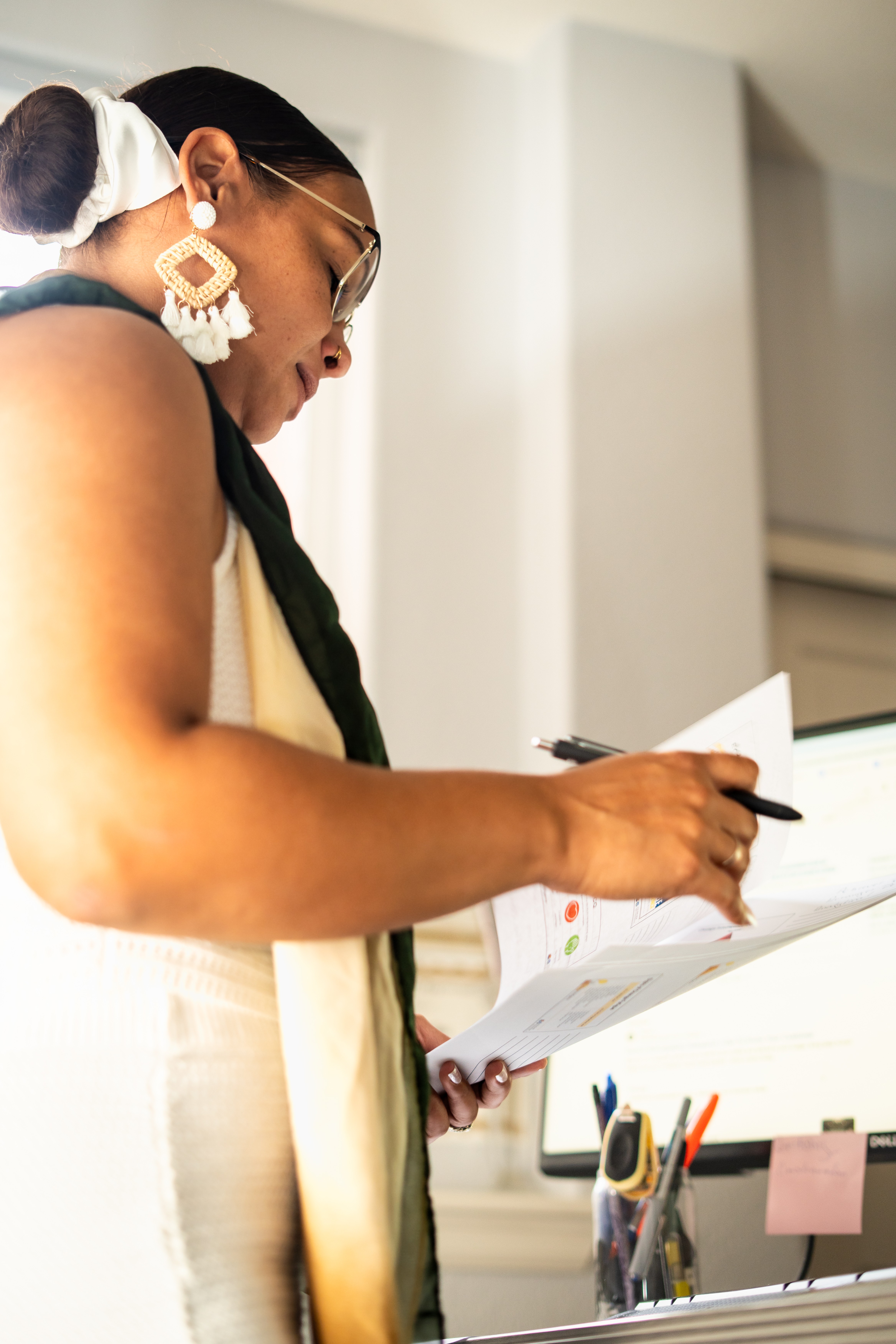 Woman with glasses and large earrings writes on paper, standing near a desk with pens and office supplies.