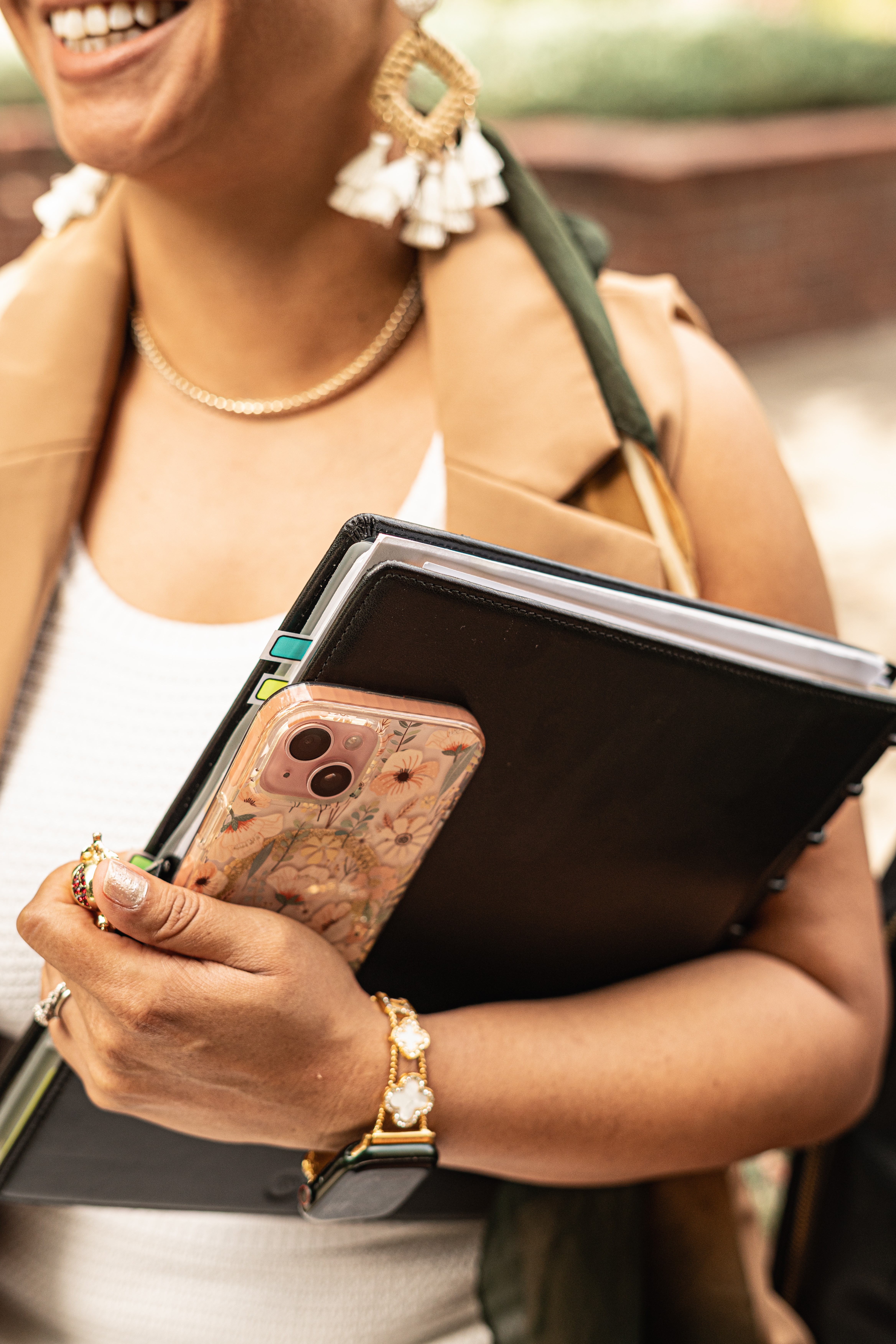 Person smiling, holding a phone and notebooks, wearing earrings, a necklace, and a watch, outdoors.