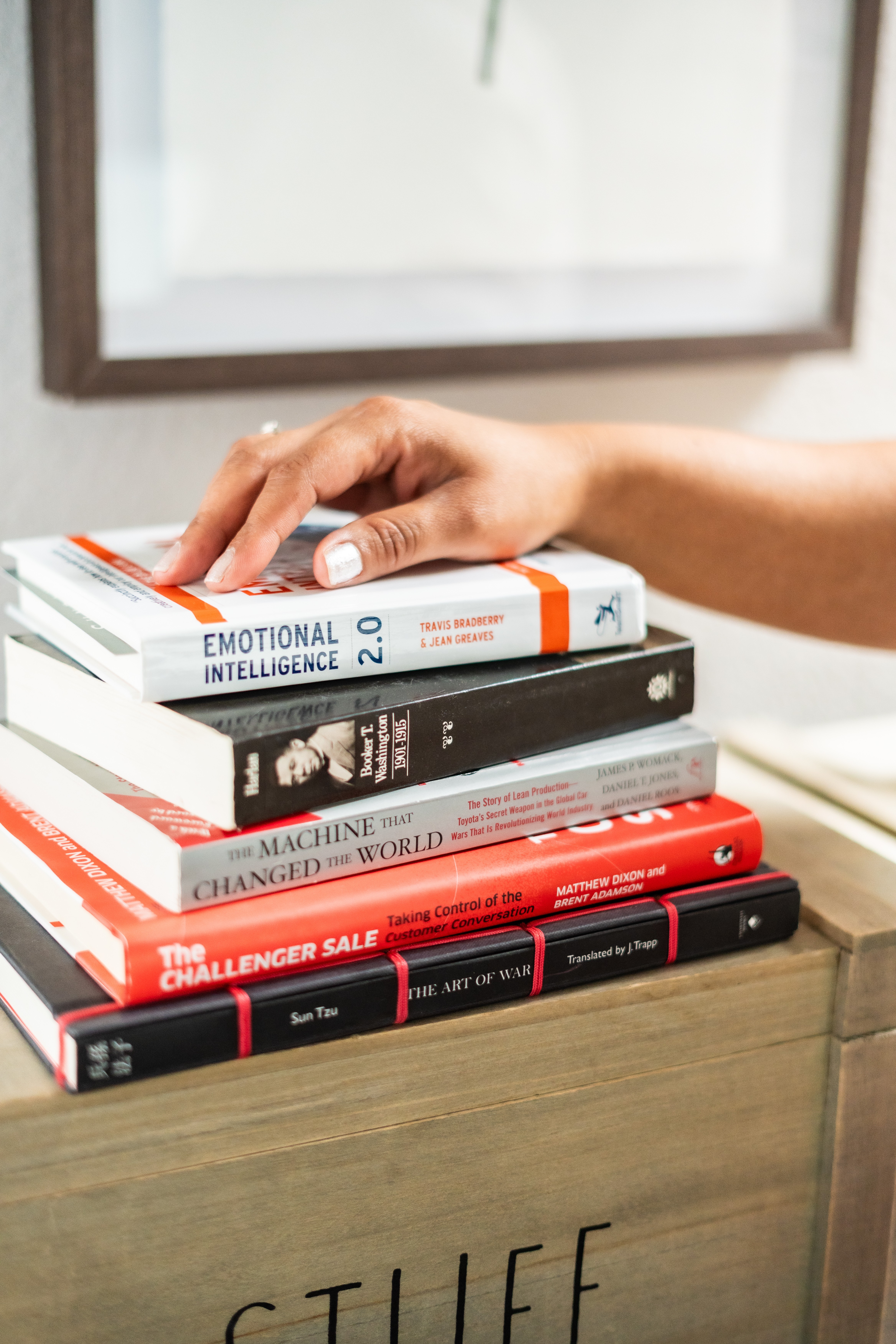 Person's hand resting on a stack of books on a wooden surface, with a framed picture in the background.