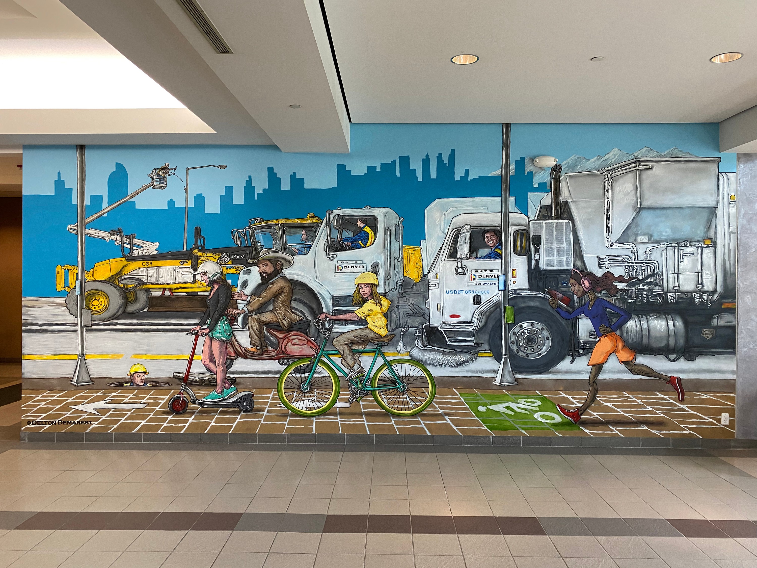 Colorful mural of cityscape with trucks, people, and a bicycle on a wall, with a sidewalk in front.