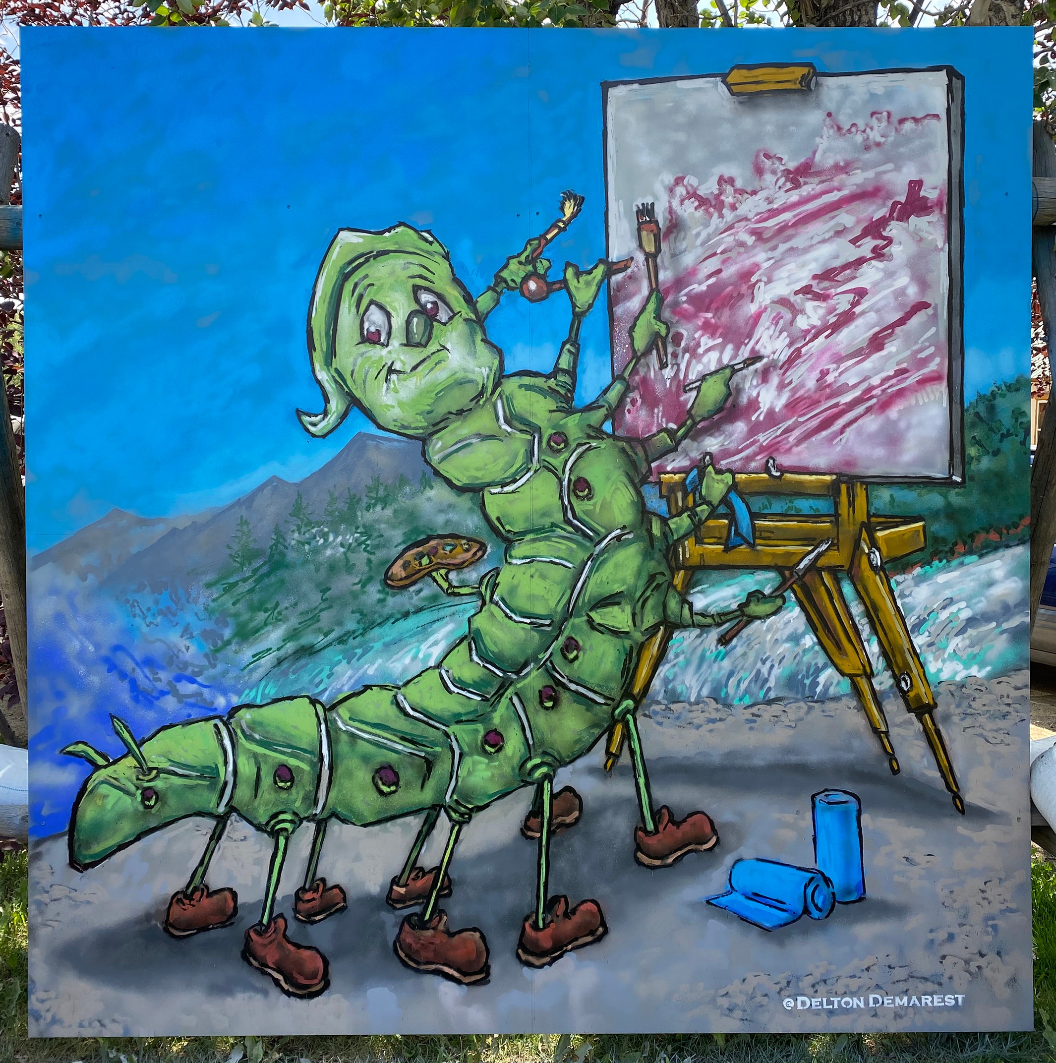 A green caterpillar with a smiling face paints on a canvas outdoors, with mountains and sky in the background.