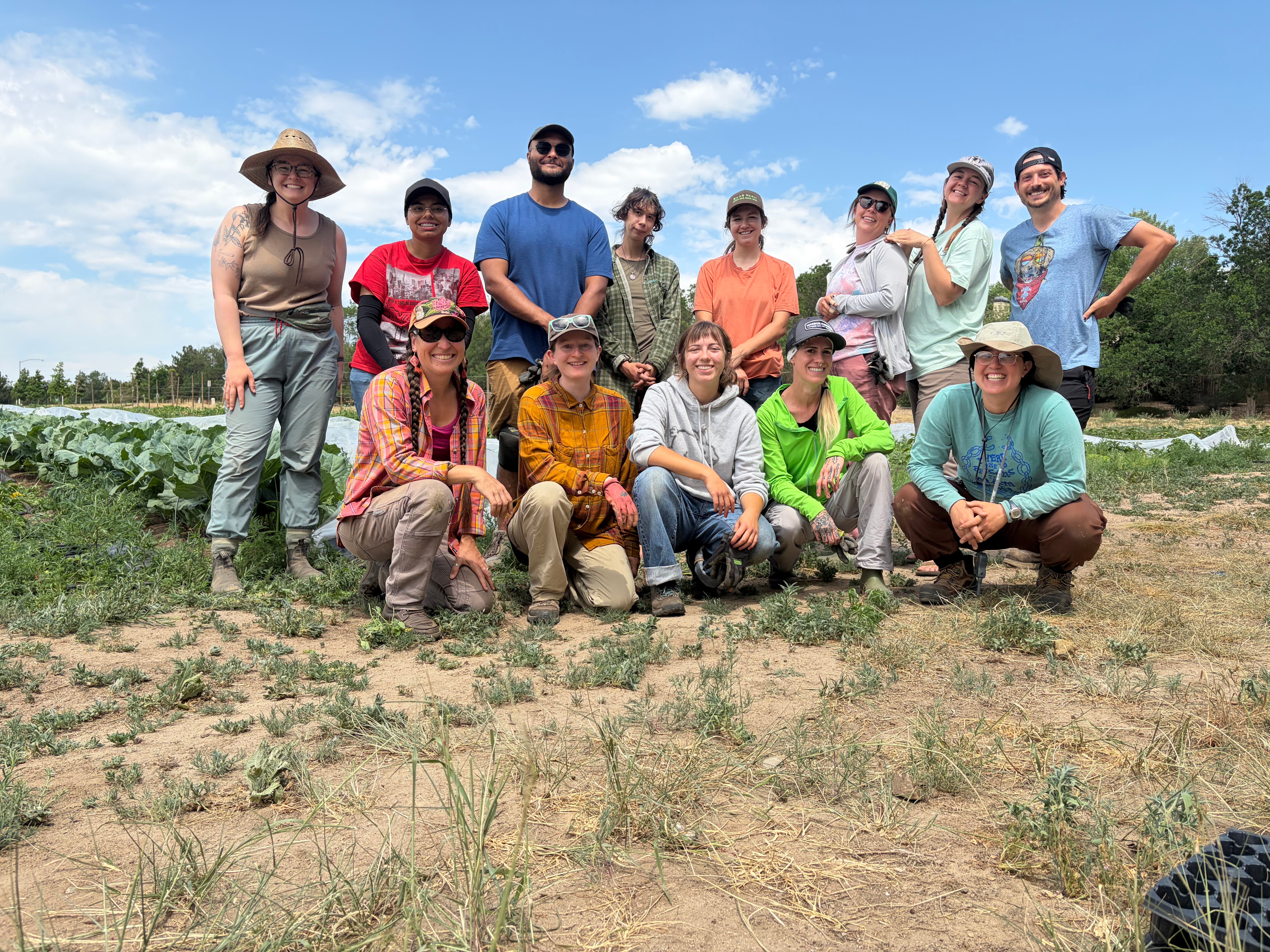 Group of people outdoors in a field, some standing and some kneeling, with blue sky and trees in background.