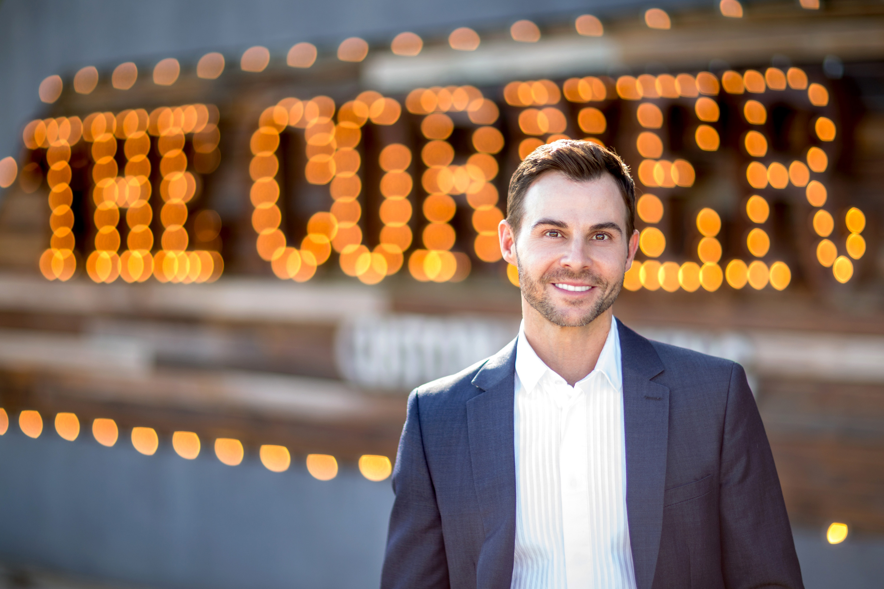Man in a suit smiling outdoors with a lit sign in the background reading 'THE CORNER'.