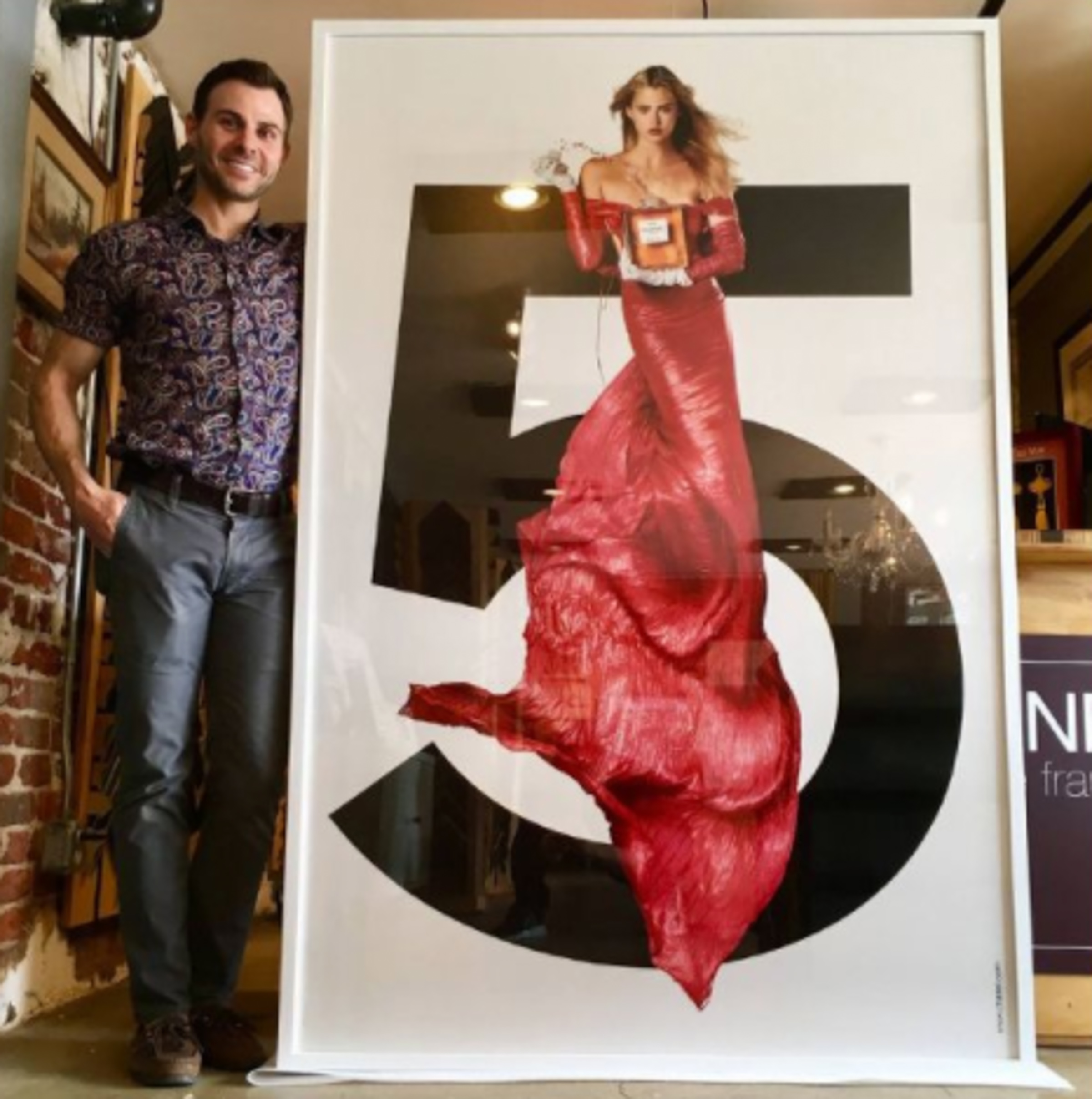 Man standing next to a large framed poster of a woman in a red gown, inside a room with artwork and furniture.