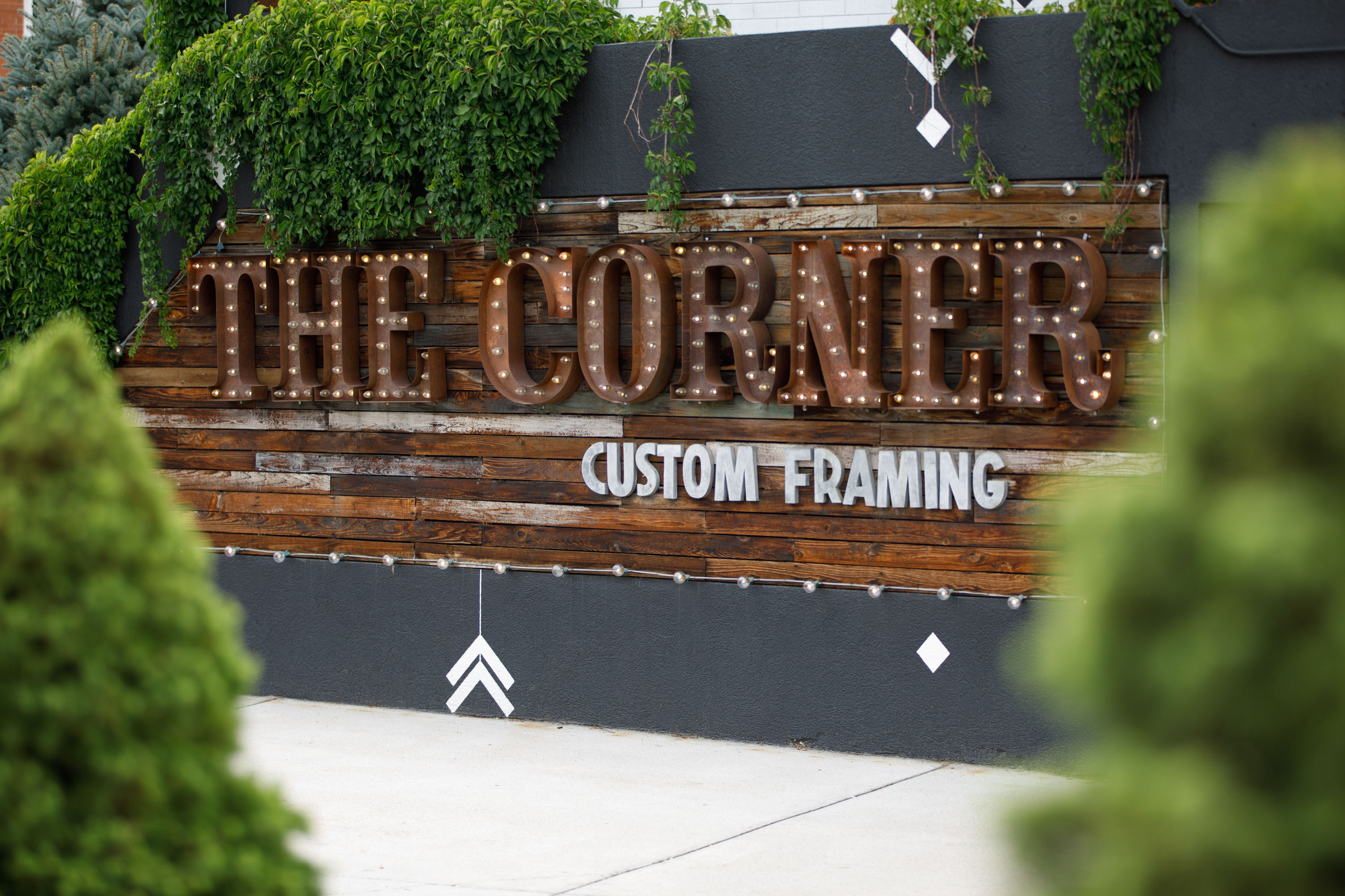 Wooden sign reads 'THE CORNER' with smaller text 'CUSTOM FRAMING' below, surrounded by greenery.