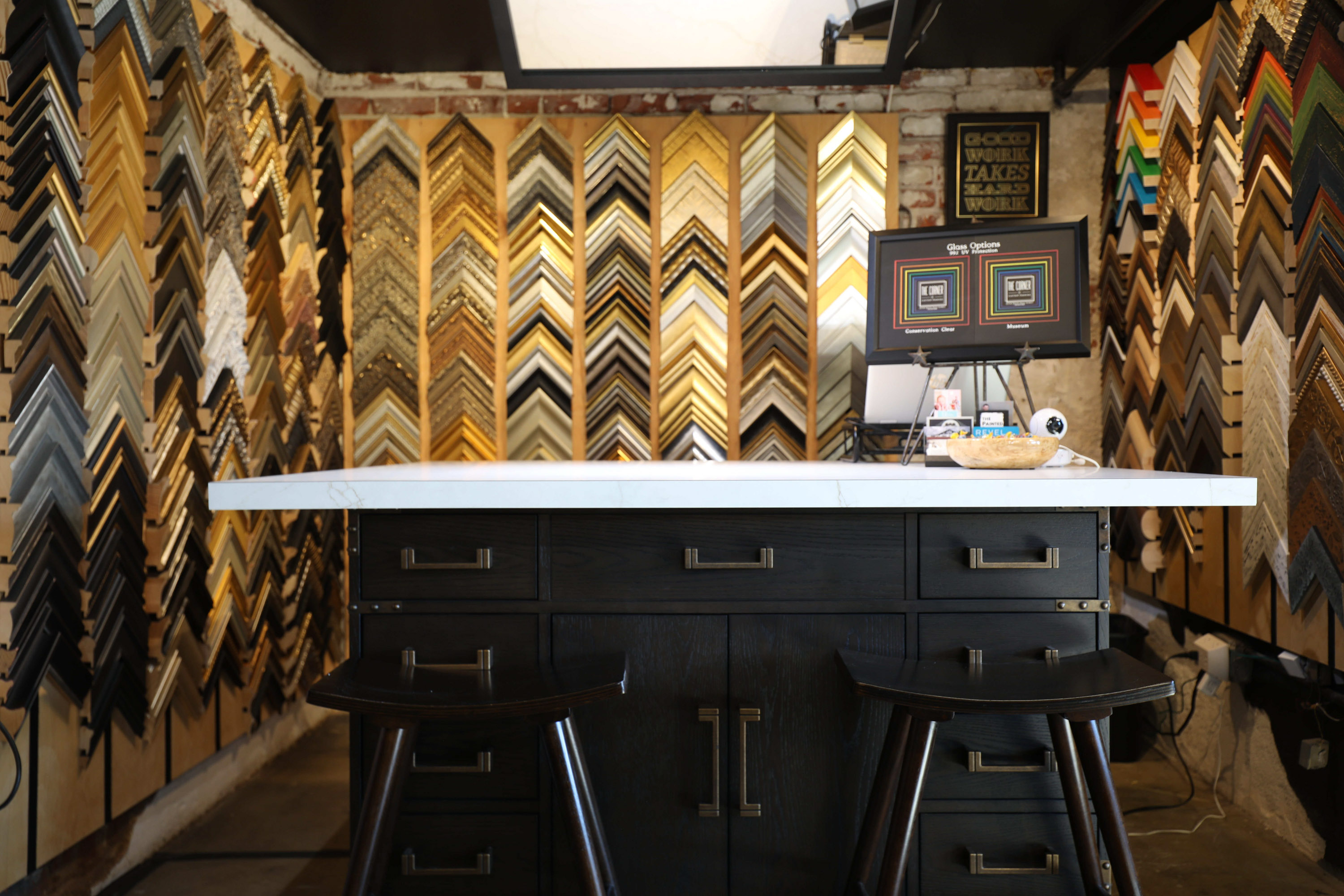 Wall covered with various picture frame samples, with a white counter and two black stools in front.