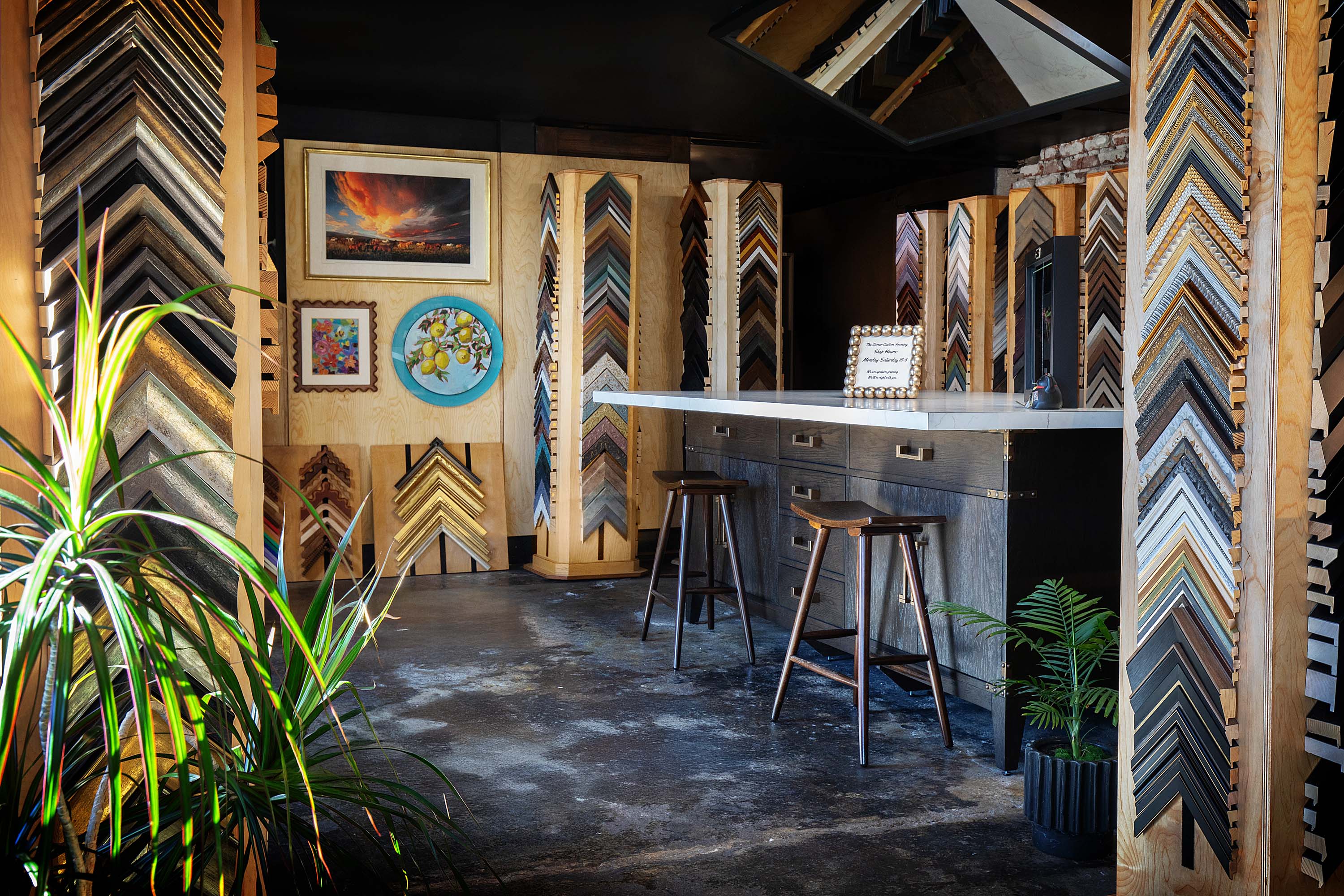 Interior space with artwork on walls, a bar counter with stools, and decorative wood paneling, plants in pots, and a dark ceiling.