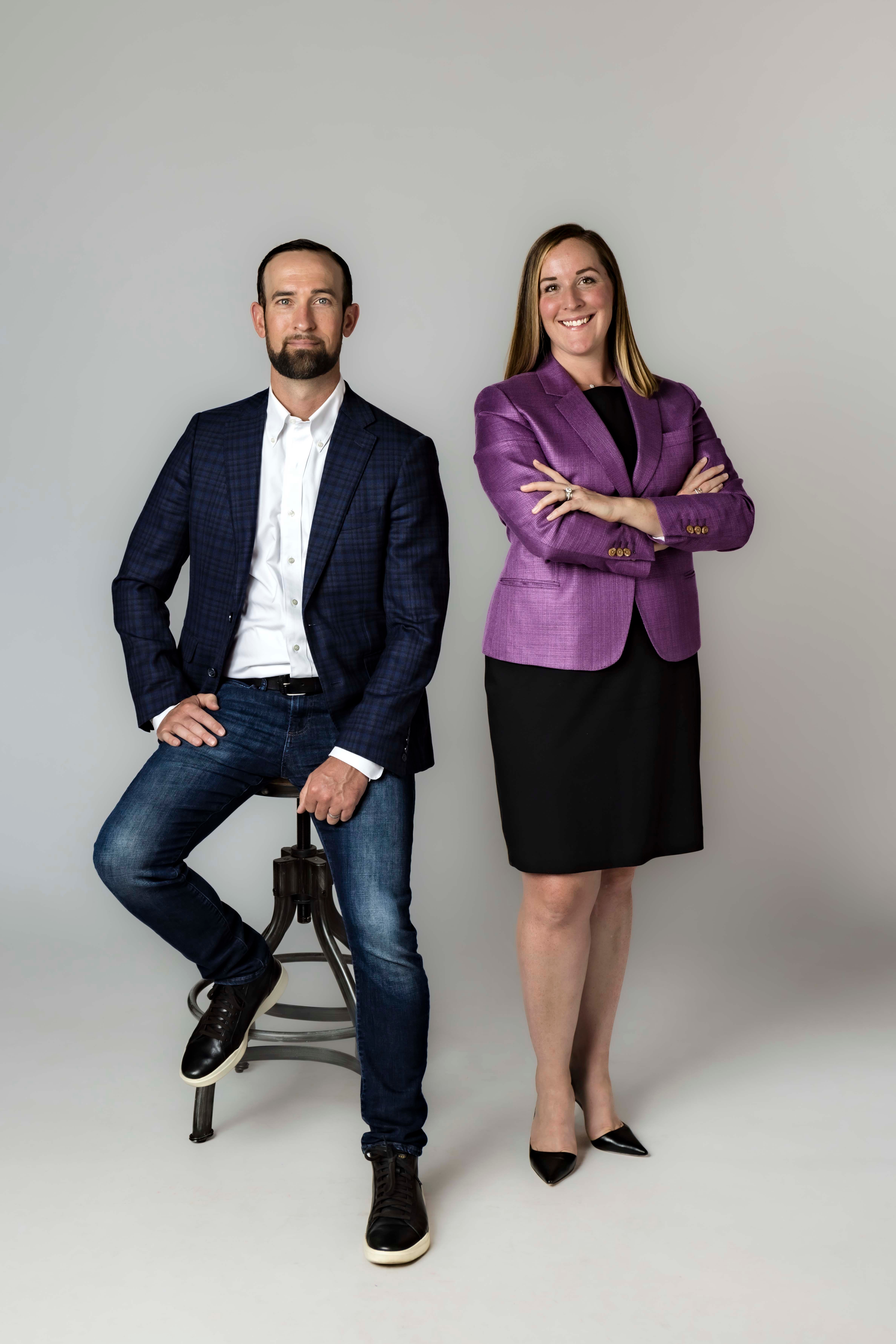 Man sitting on a stool and woman standing with arms crossed, both smiling, against a plain background.