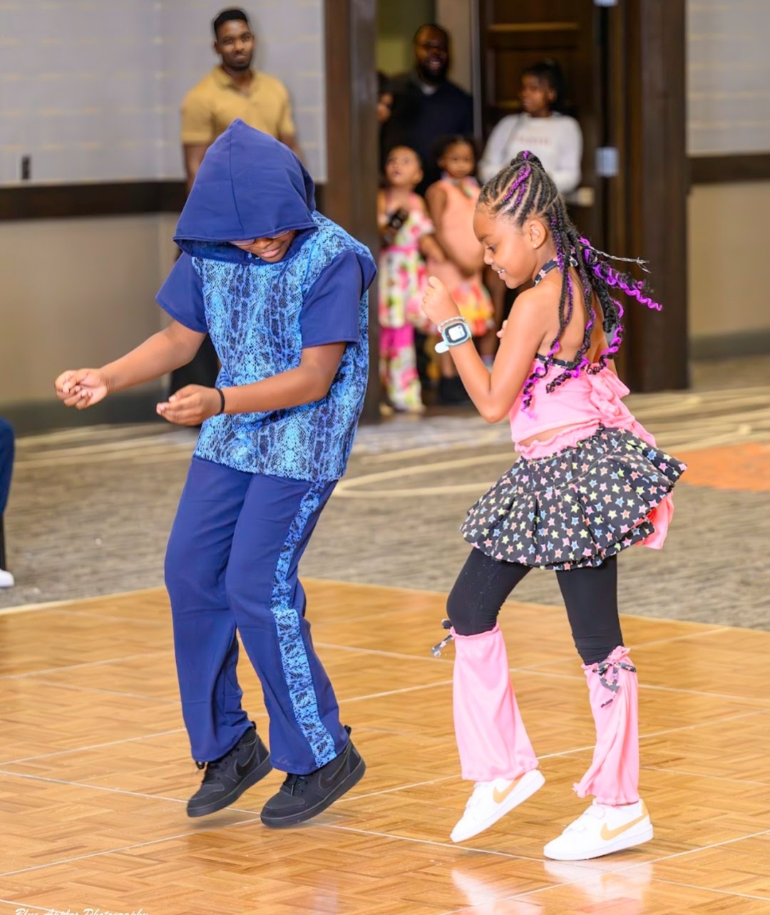 Two children dancing on a wooden floor, with onlookers in the background, one wearing a blue hoodie and the other in pink and black clothing.