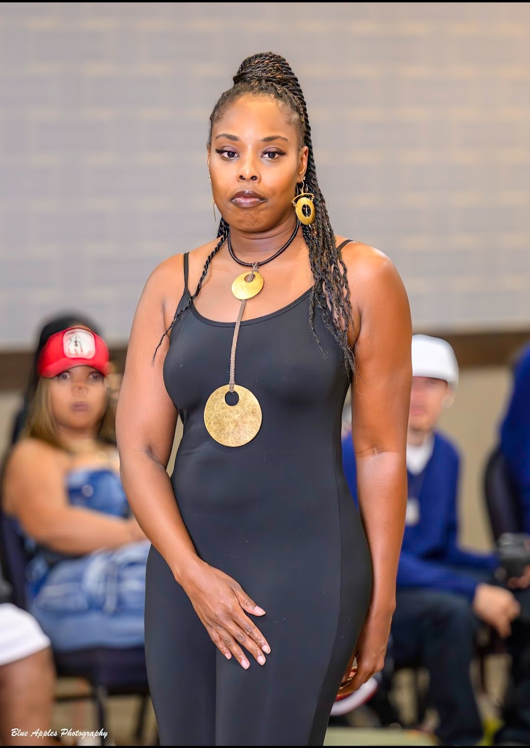 Woman with braided hair and large jewelry, wearing a black dress, standing with hands clasped, in front of seated children.