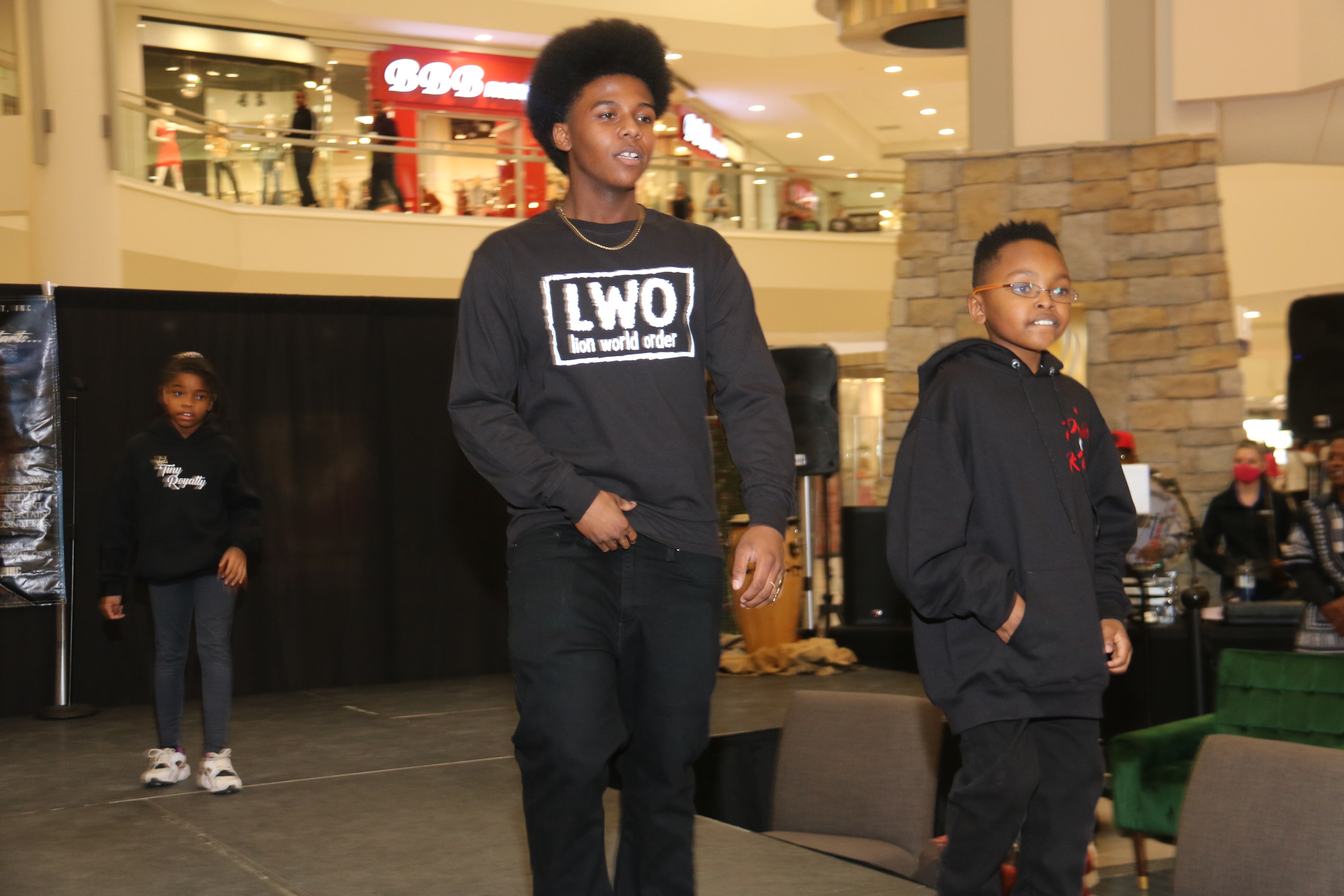 Two young people standing in a shopping mall, one with curly hair wearing a black hoodie, the other with glasses and a black jacket.