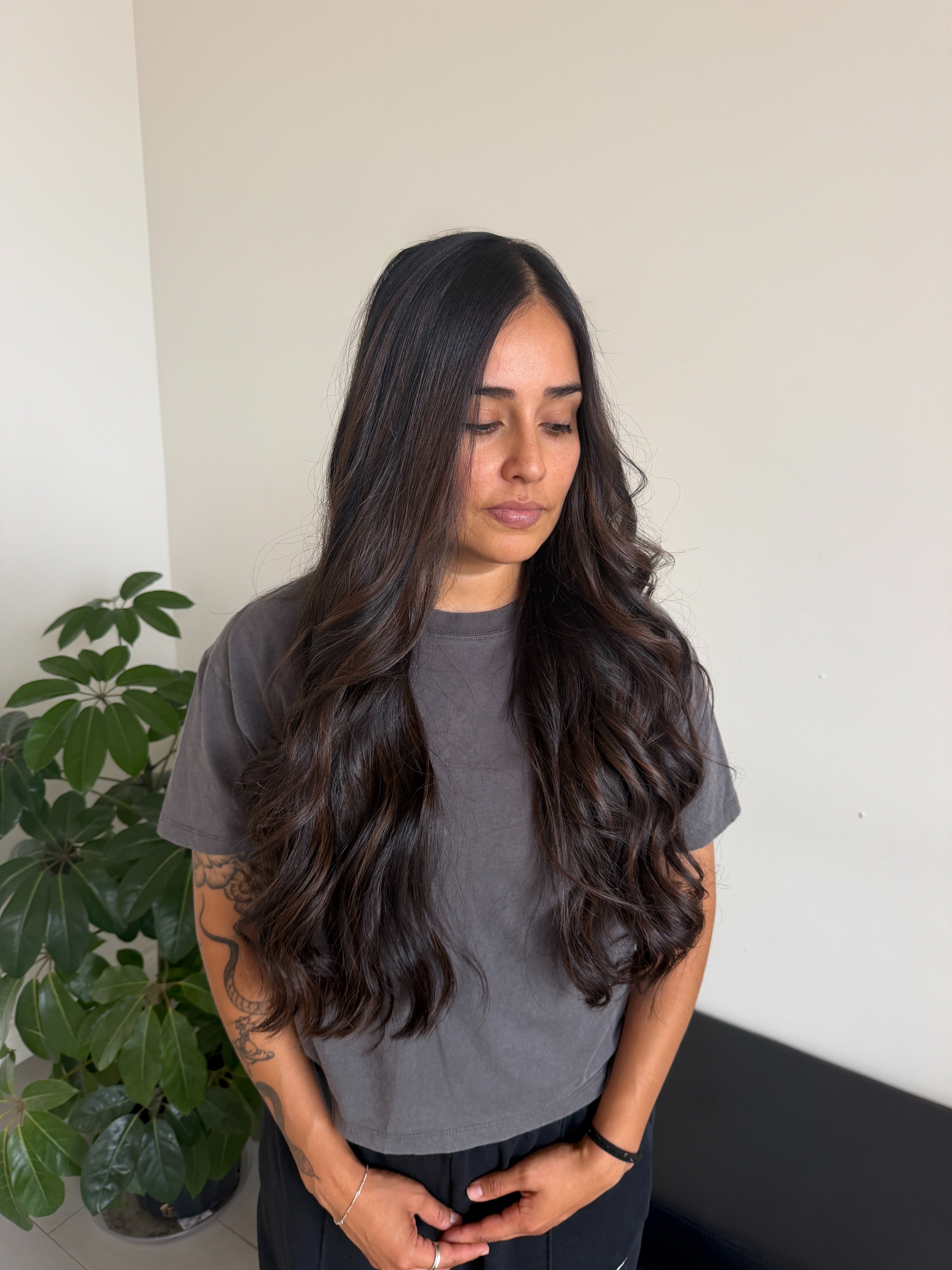 Young woman with long wavy dark hair wearing a gray t-shirt, standing indoors near a plant, looking down.