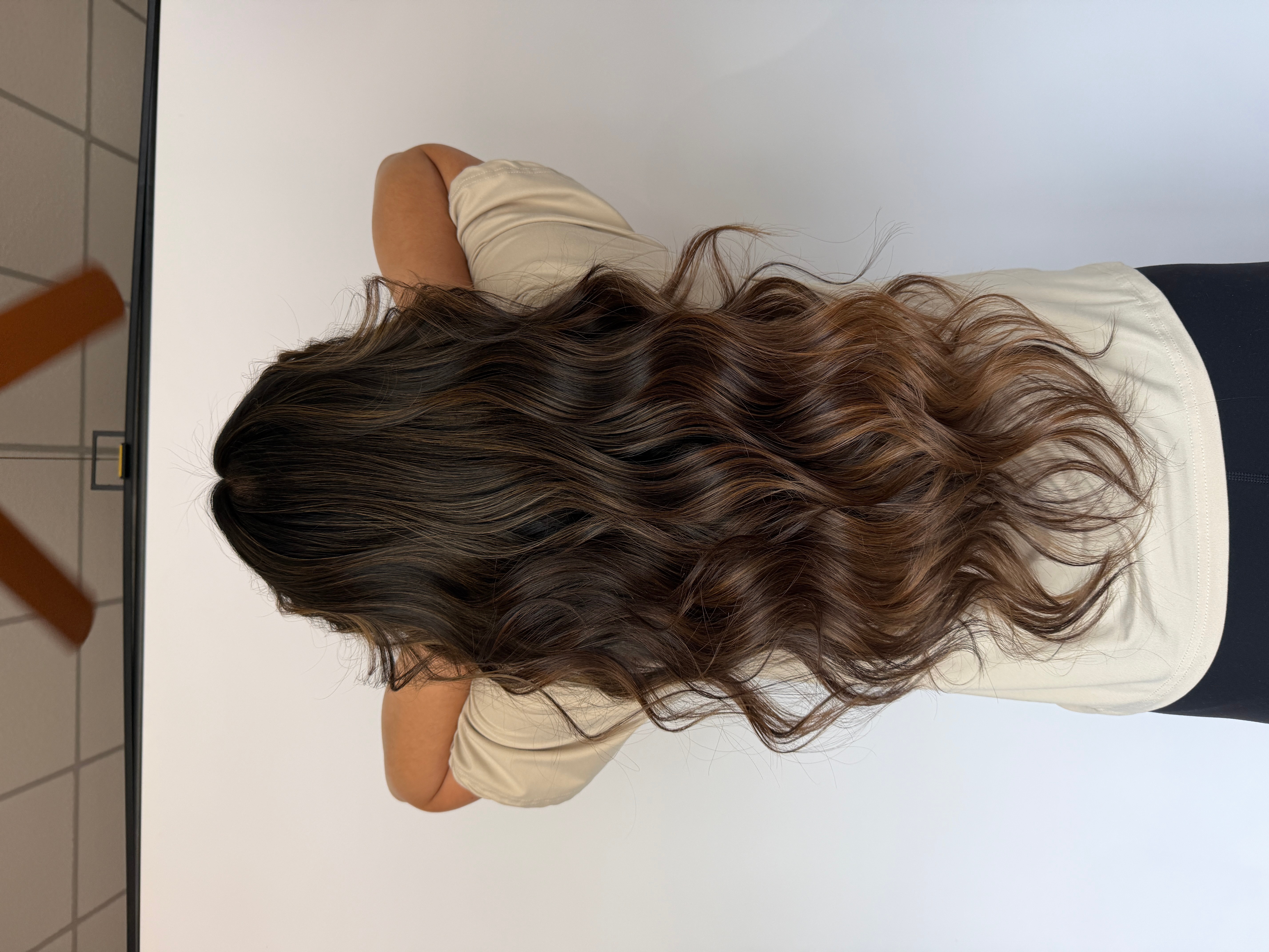 Person with long, wavy hair facing away, resting arm on a surface, wearing a light-colored shirt, in a neutral setting.