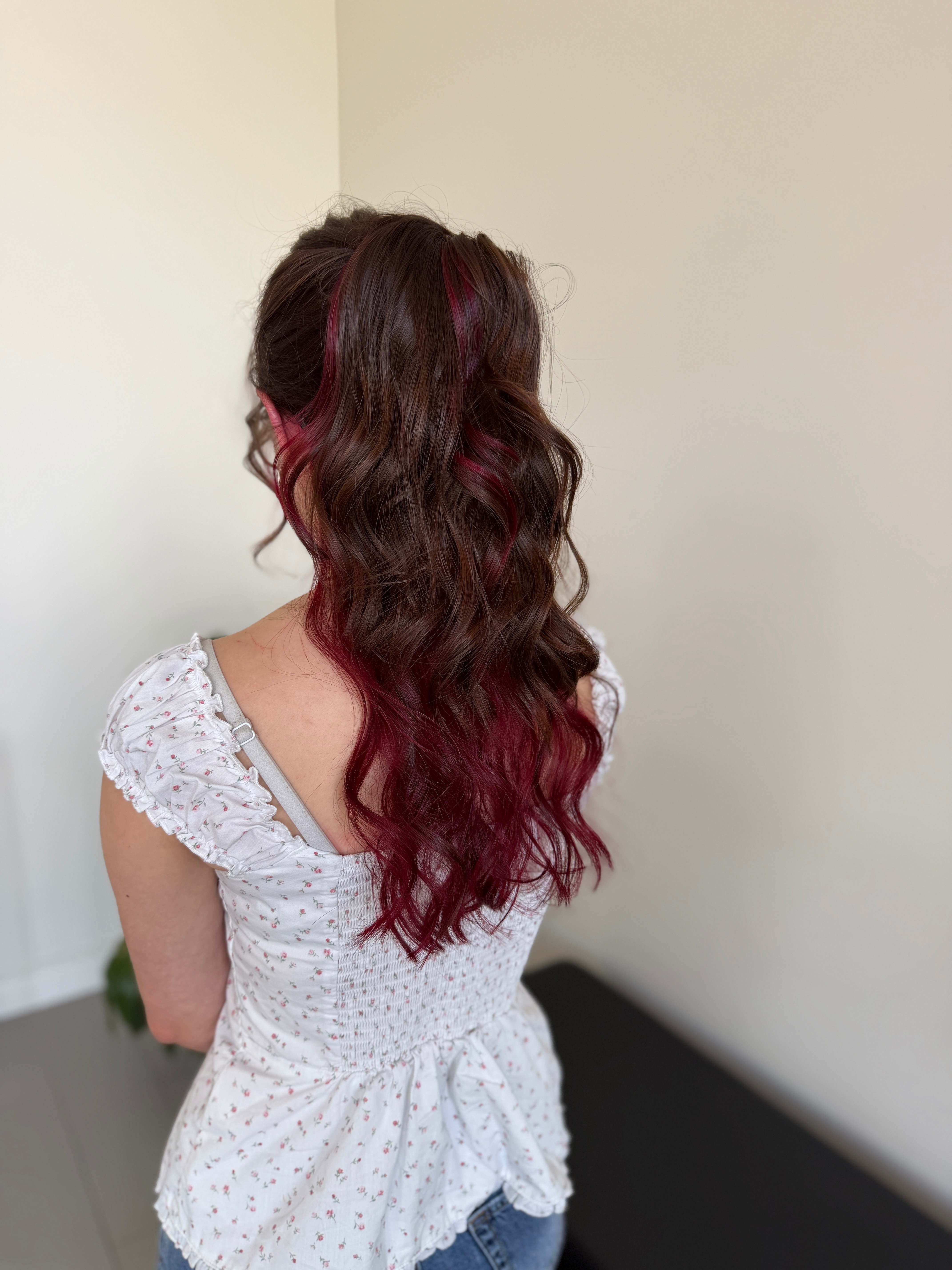 Person with long, wavy hair dyed in shades of red and pink, wearing a white, short-sleeved, patterned dress, facing away from the camera.