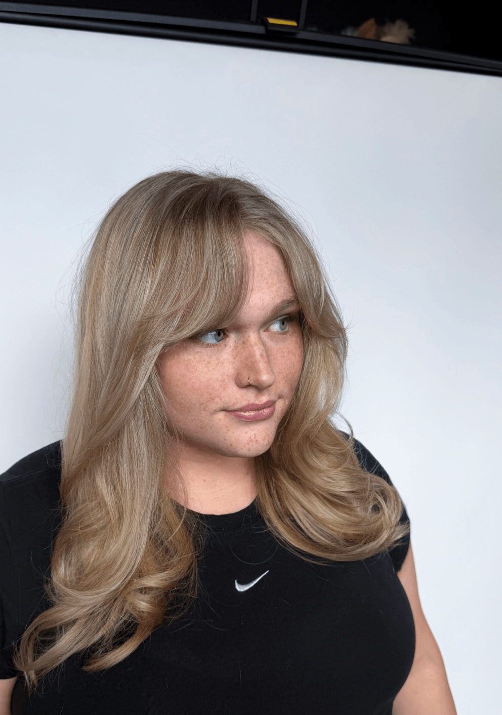 Woman with long blonde hair and black shirt looking to the side, against a plain background.