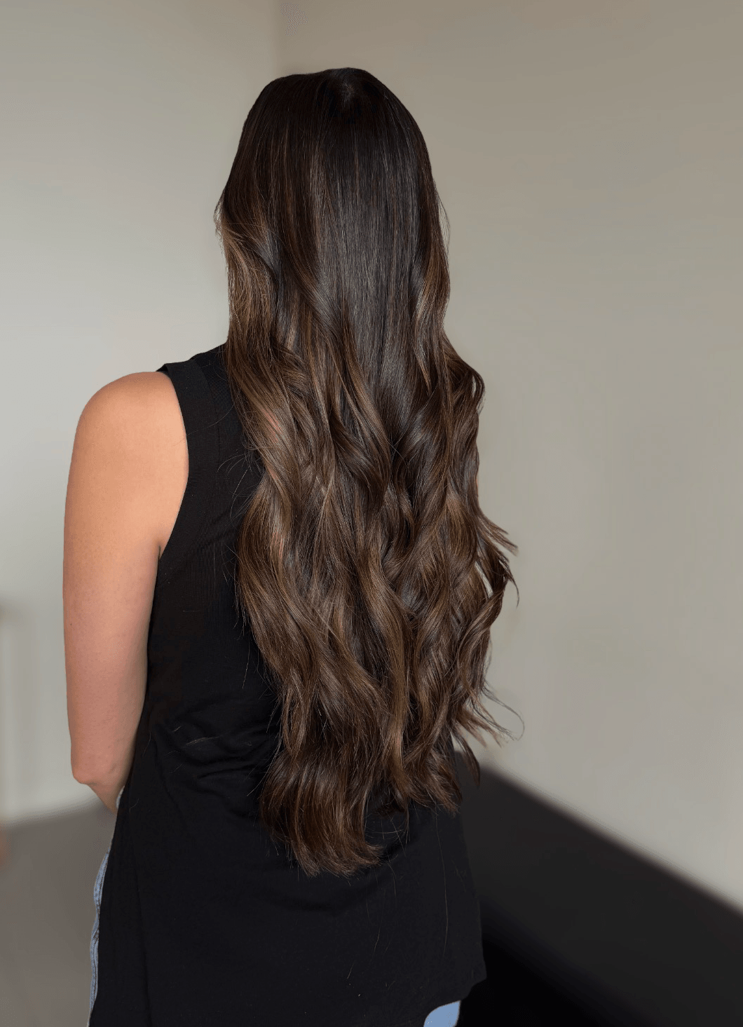 Woman with long, wavy brown hair wearing a black sleeveless top, facing away from the camera.