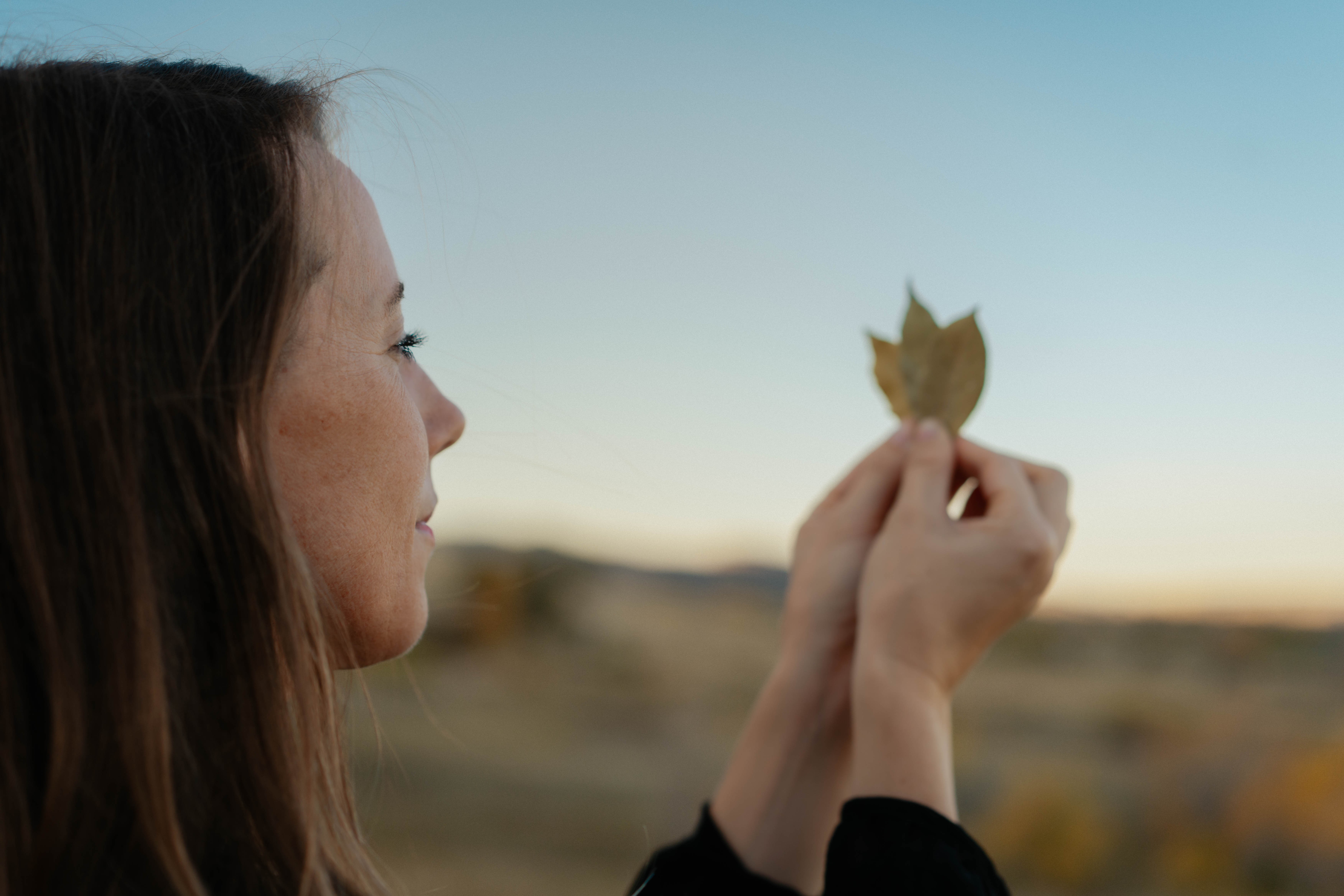 Woman holding a leaf outdoors during sunset, with a blurred landscape background.