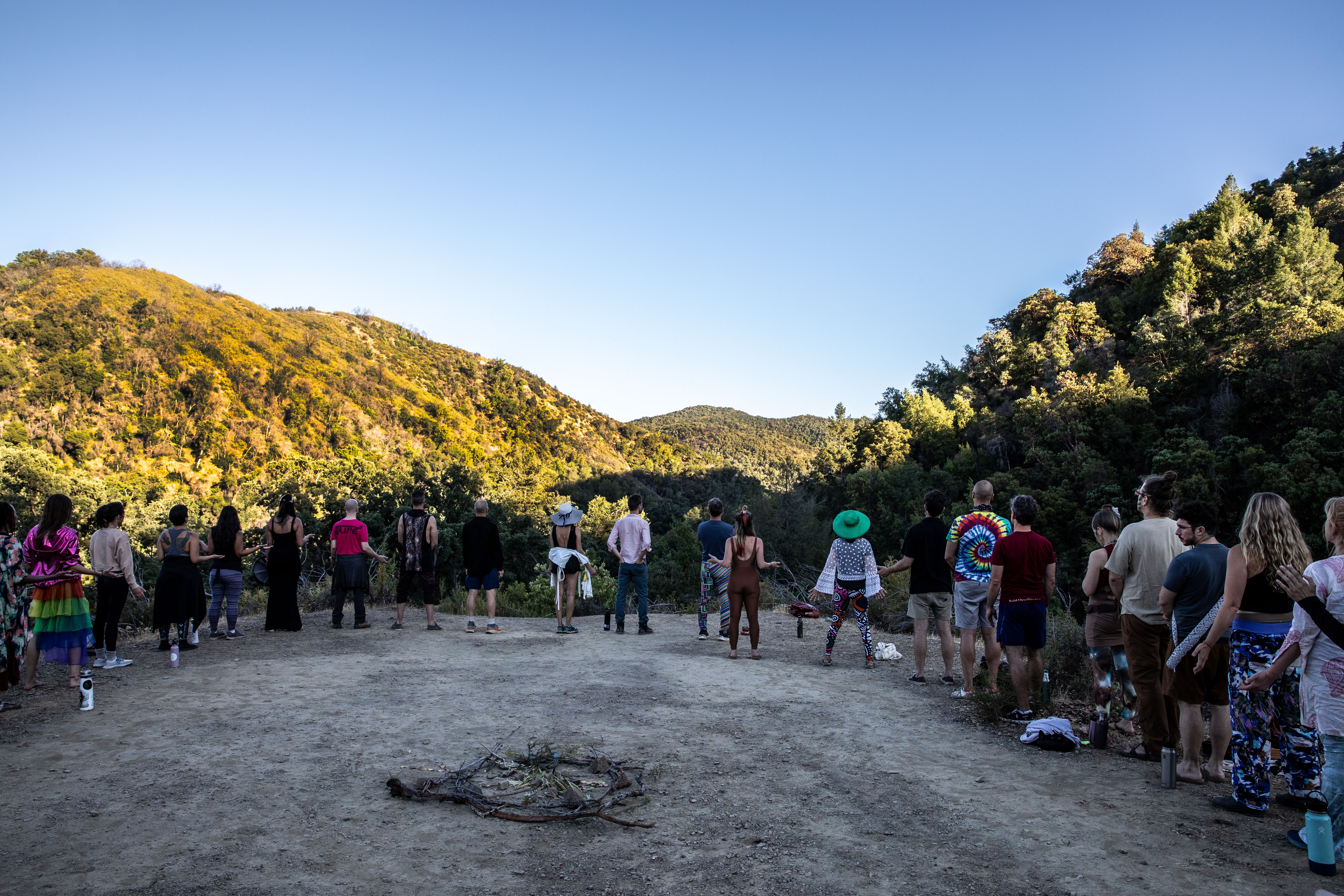 Group of people standing on a dirt path in a hilly, forested area under a clear blue sky, some taking photos.