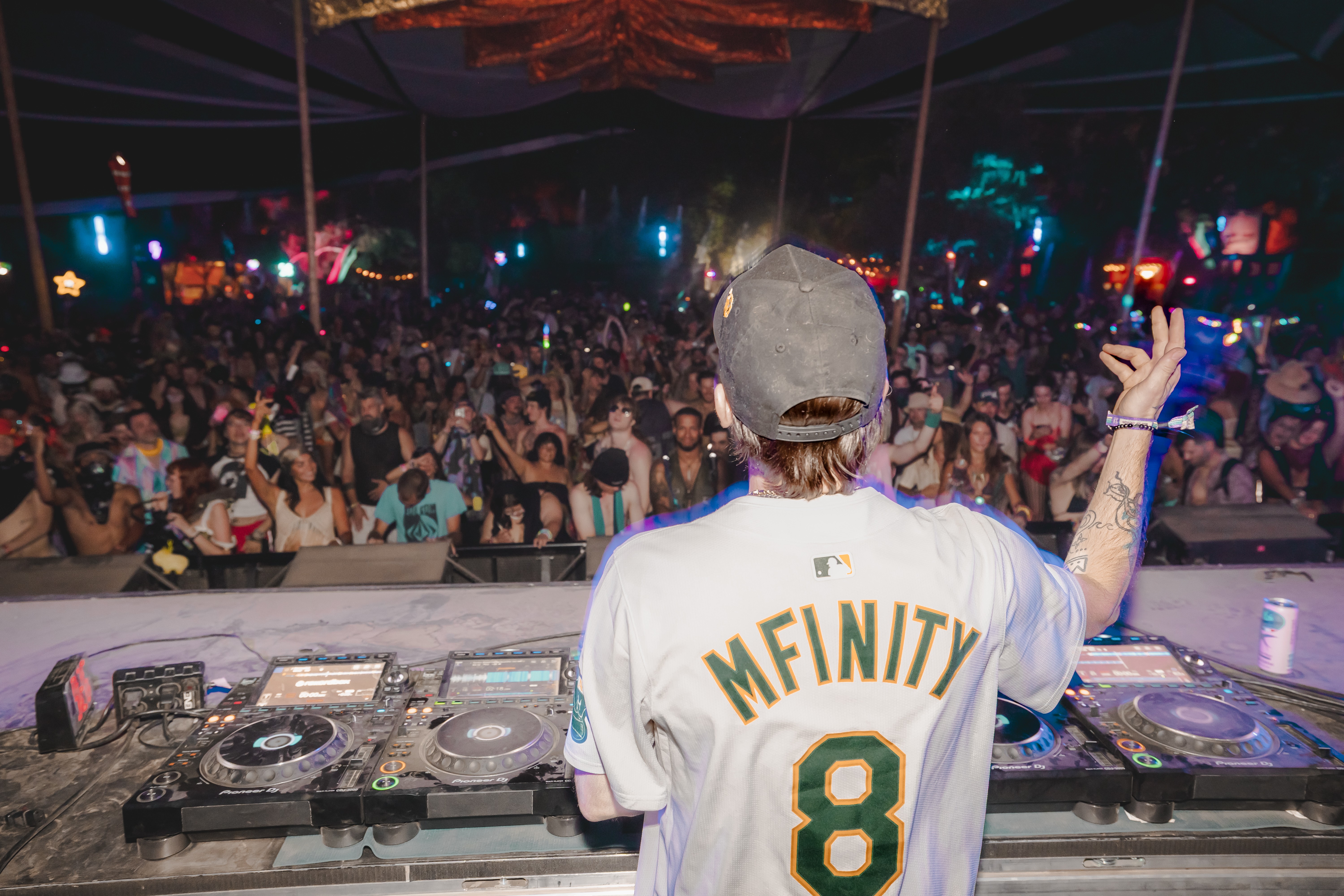 DJ performing at a crowded outdoor music event at night, with a large audience and colorful lights in the background.