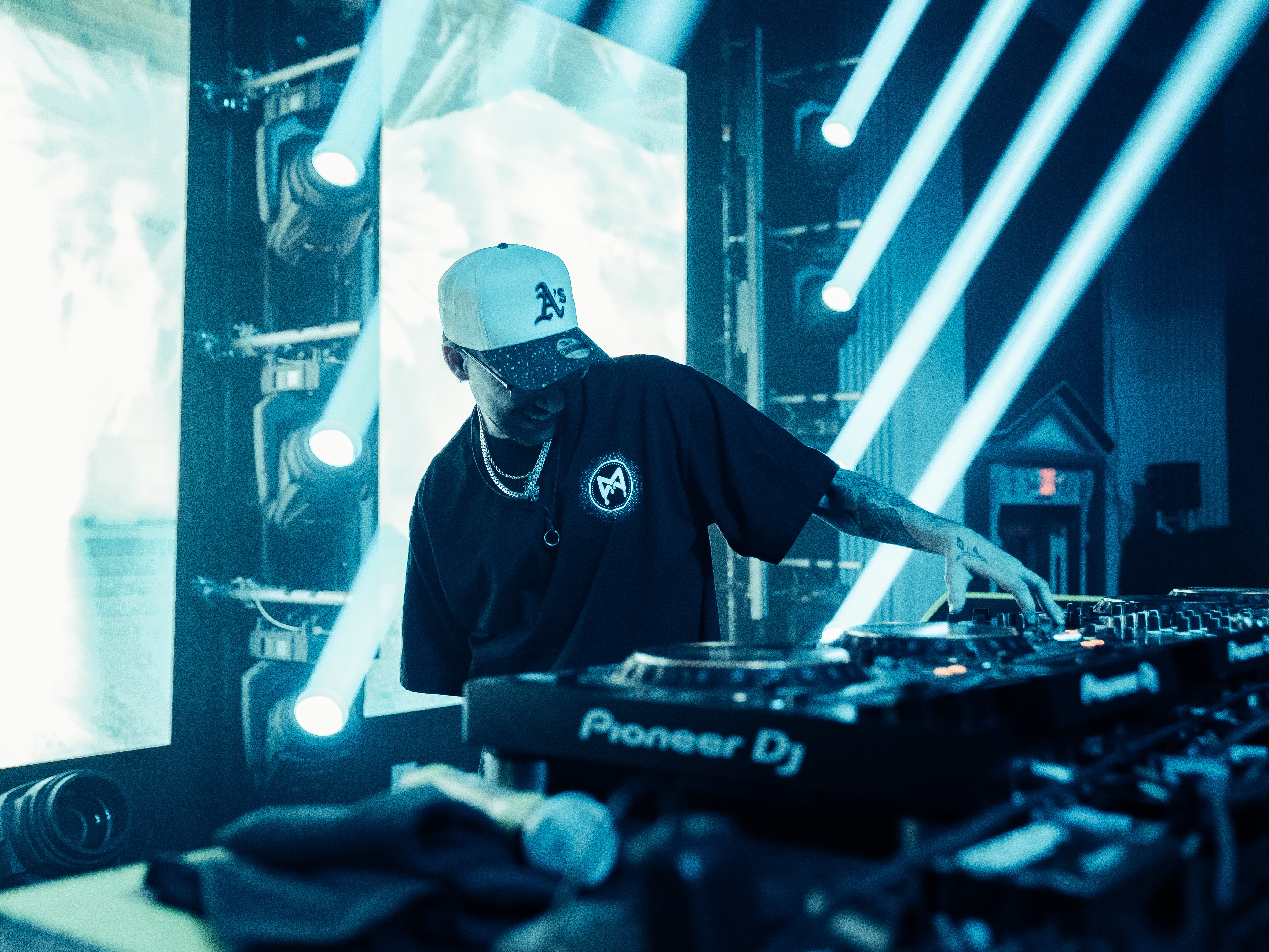 DJ wearing a cap and black shirt, operating equipment on stage with bright lights and screens in background.