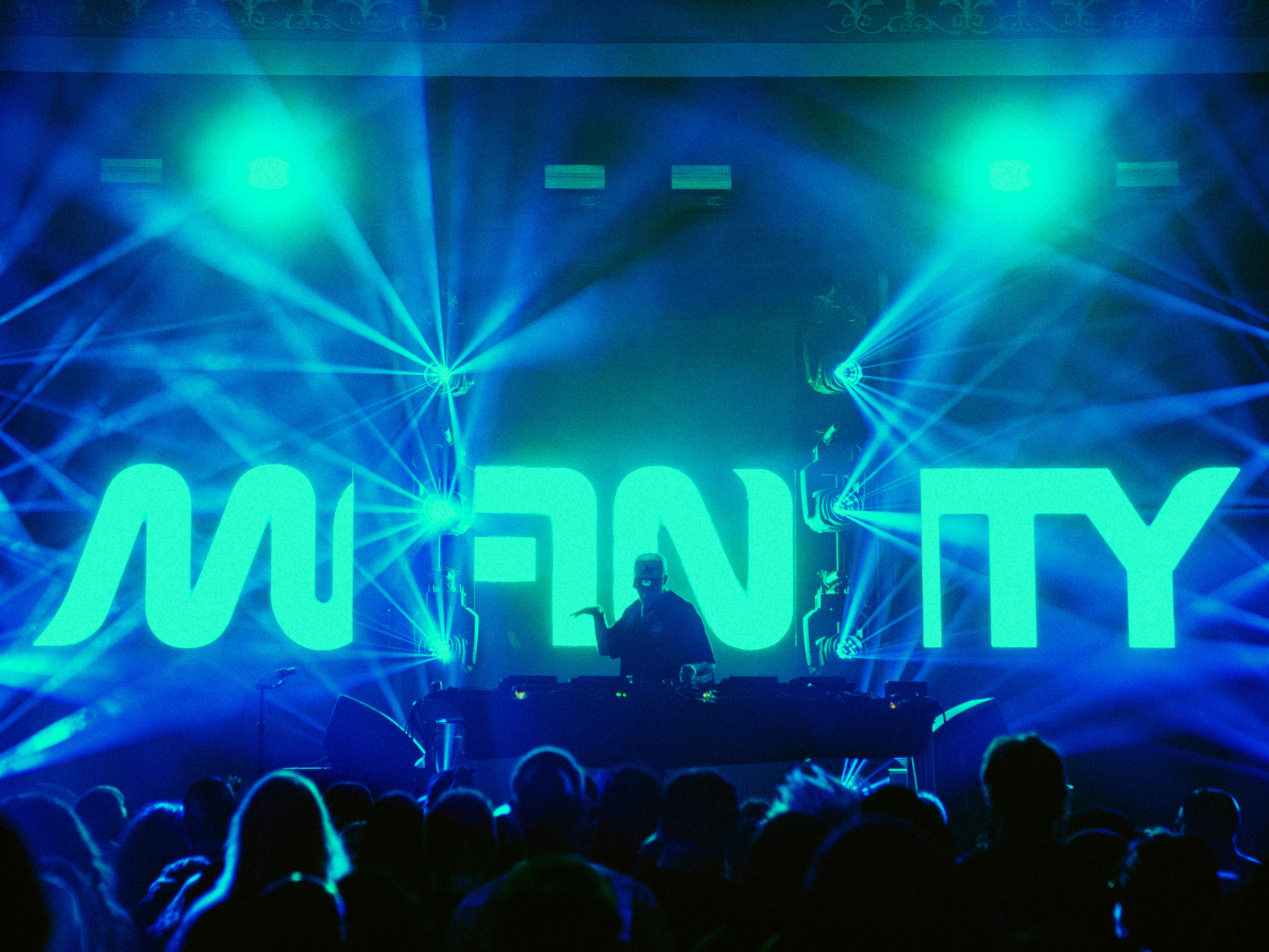 Stage with bright blue and green lights, large illuminated text, and a DJ performing in front of an audience.