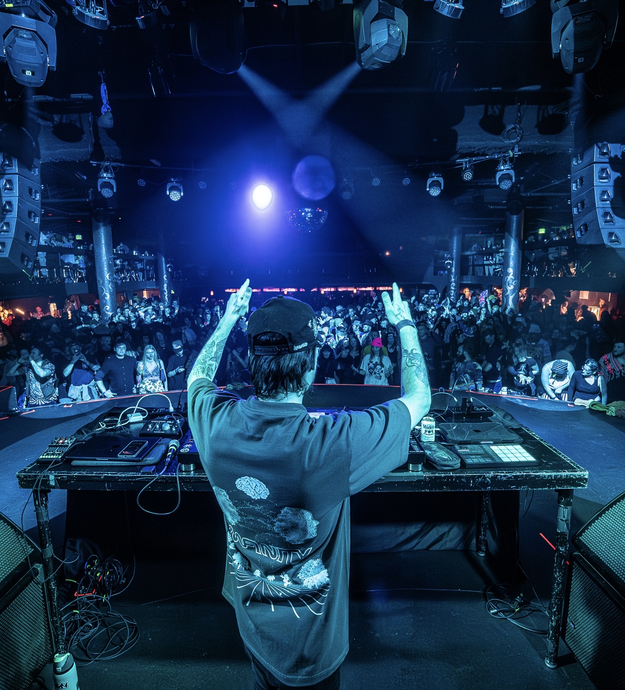 DJ performing at a nightclub with a large crowd, blue lighting, and sound equipment visible, viewed from behind the DJ.
