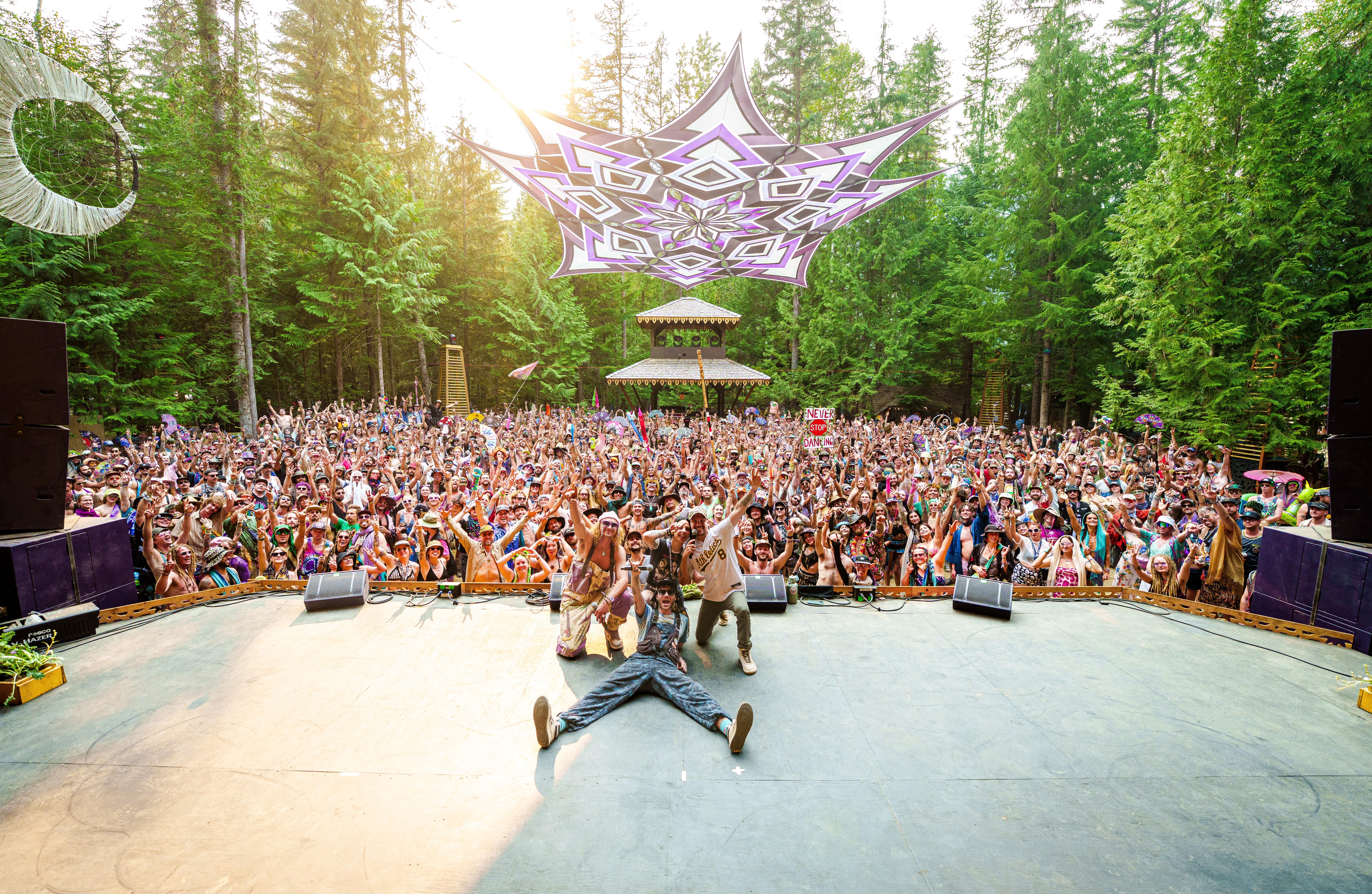 Performer lying on stage with a large crowd and forest background, colorful canopy overhead, outdoor concert scene.