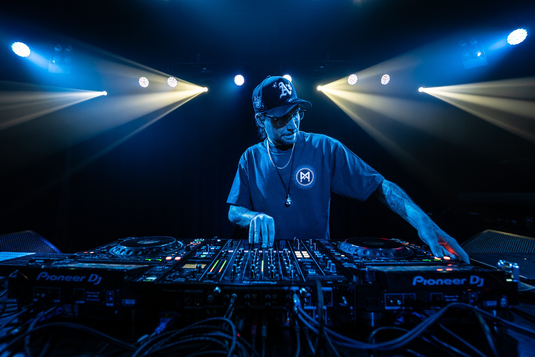 DJ wearing a cap and mask, operating turntables with colorful stage lights behind, in a dark setting.