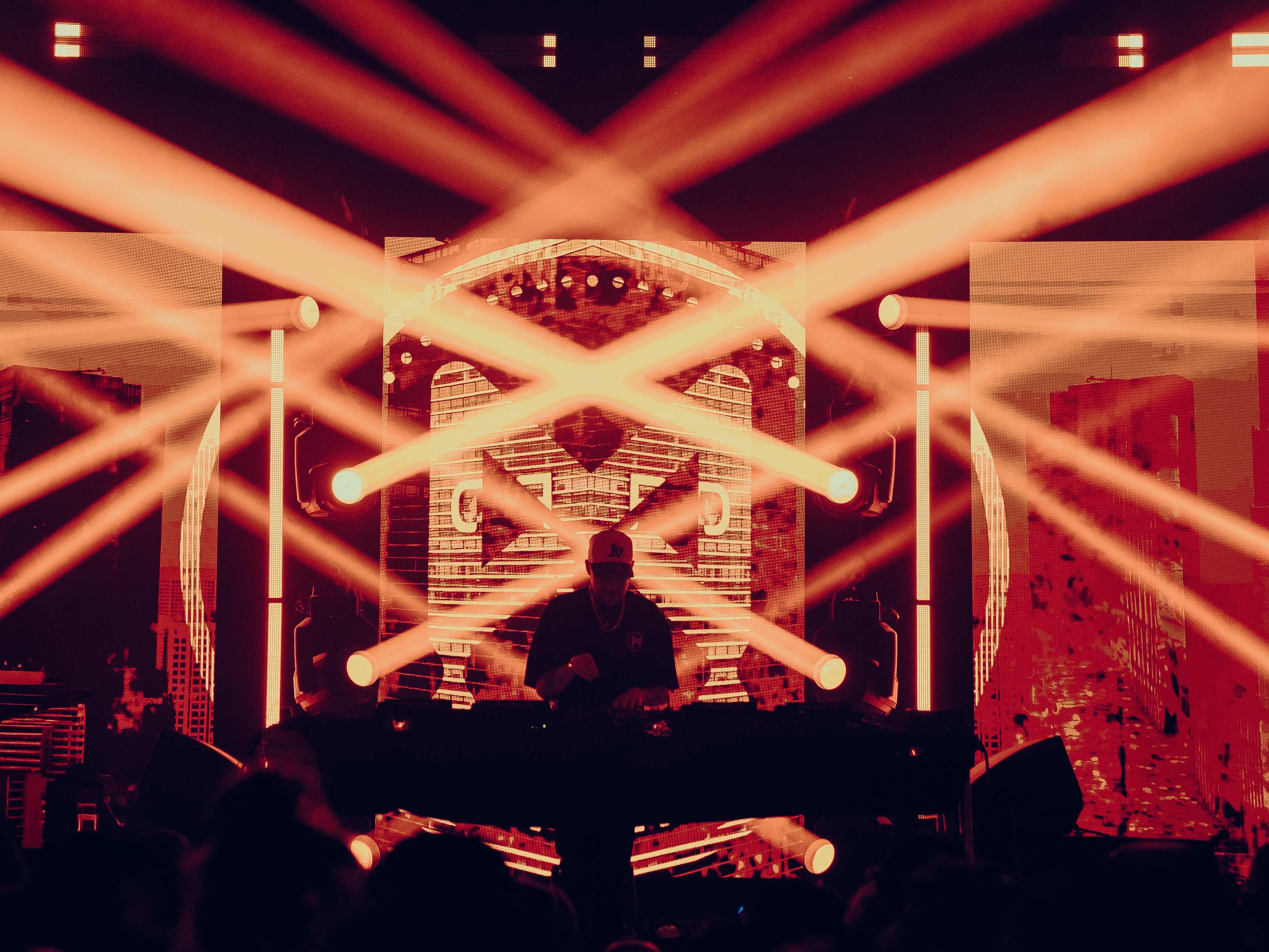 DJ performing on stage with bright orange and yellow light beams, heart-shaped decoration, and audience silhouettes in foreground.
