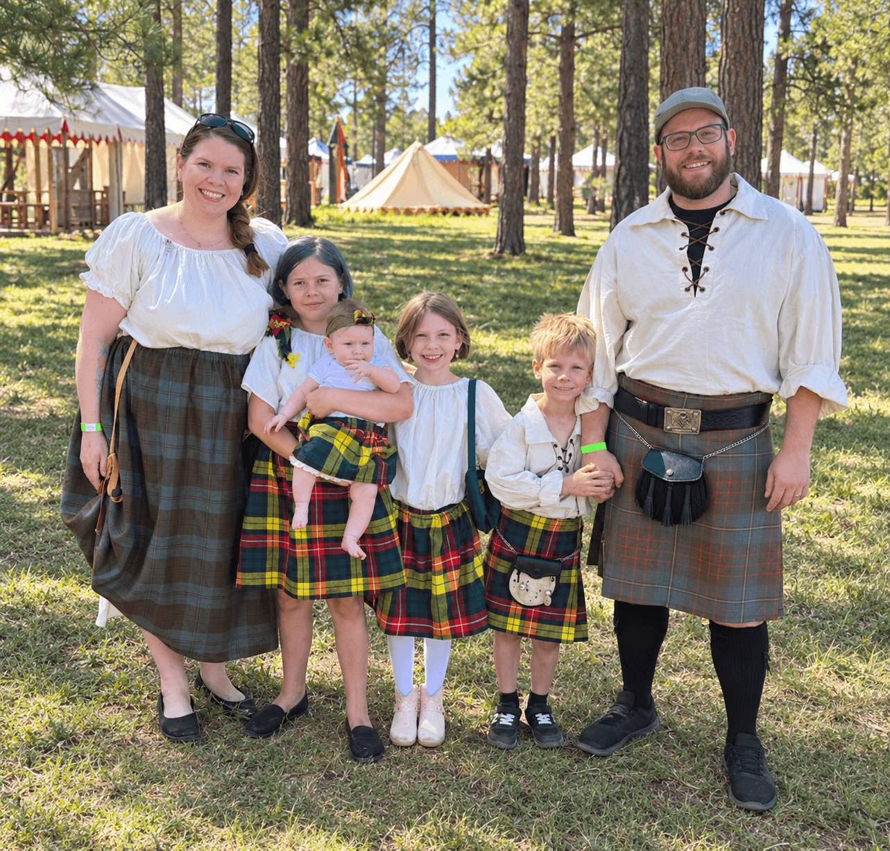 Family of six in traditional clothing outdoors, trees and tents in background, smiling at camera.