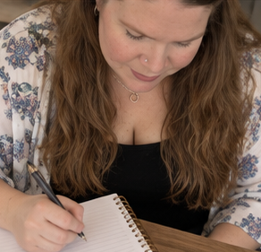 Woman with long hair writing in a notebook, wearing a patterned top and black shirt, looking down.