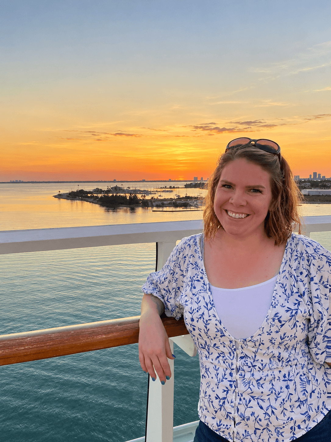 Smiling woman with sunglasses on her head leaning on a railing during sunset over water.