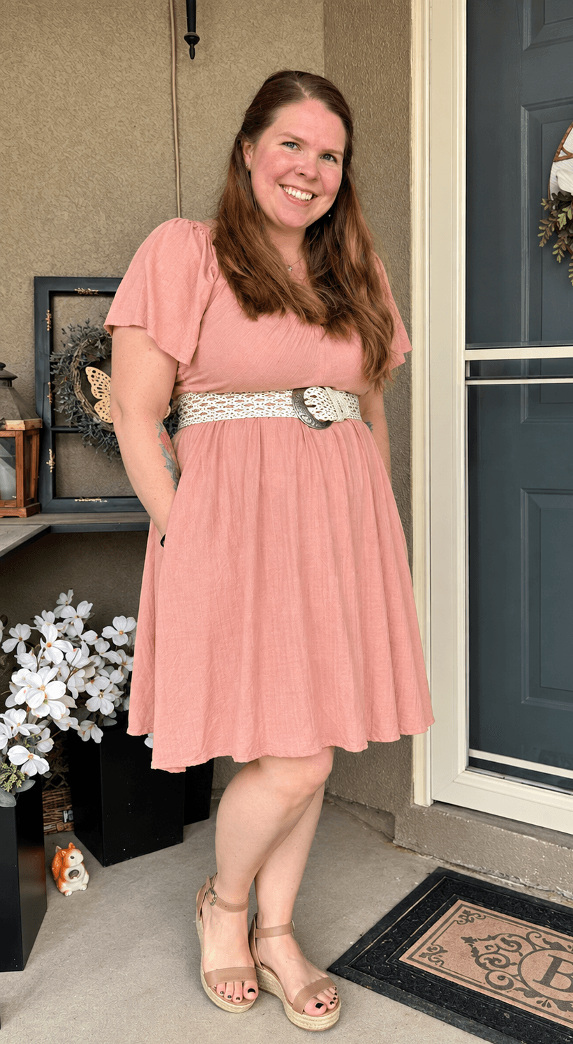 Woman in pink dress standing outside near a door, smiling, with flowers nearby.