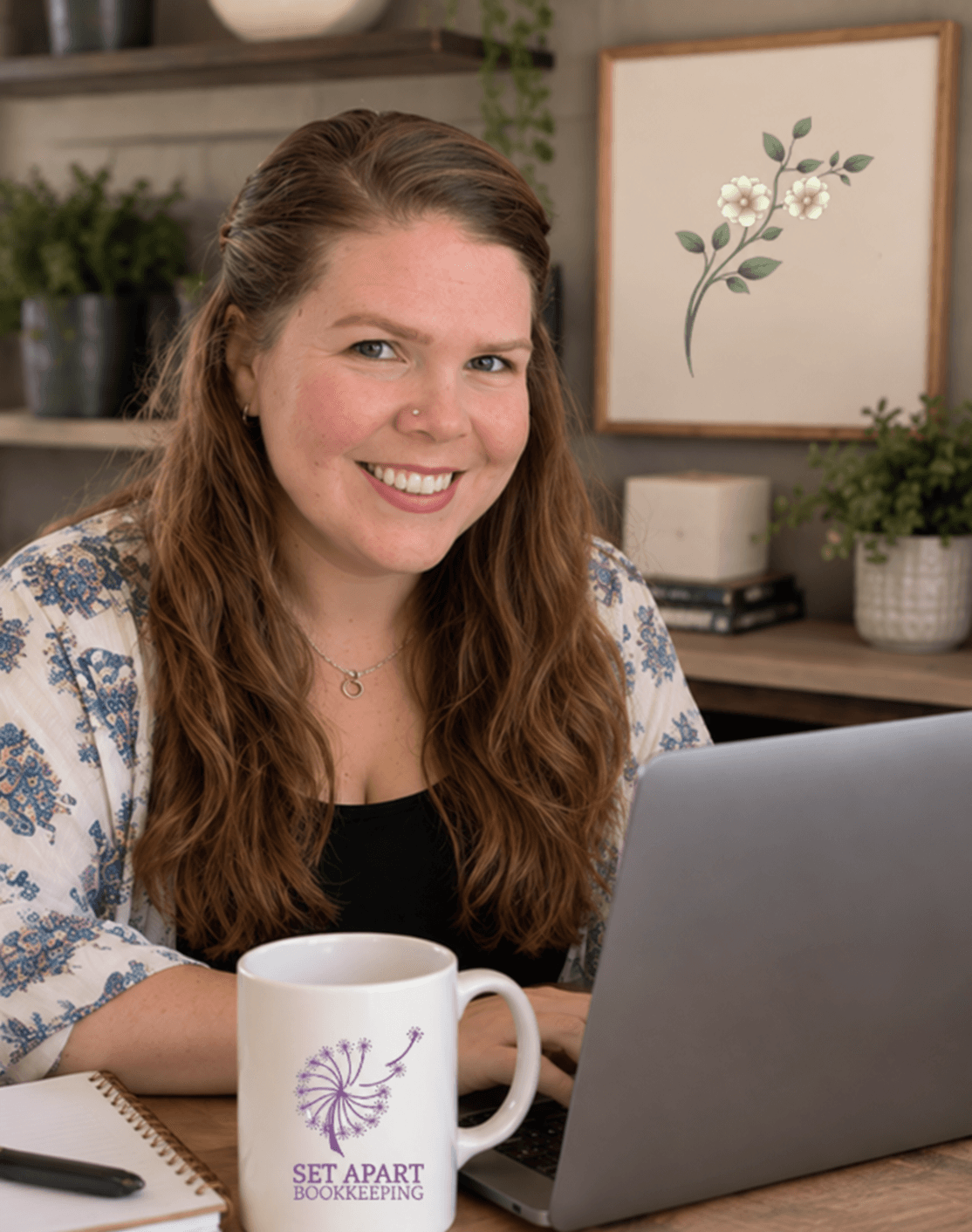 Woman with long wavy hair smiling at a laptop, with a mug and plants in background.