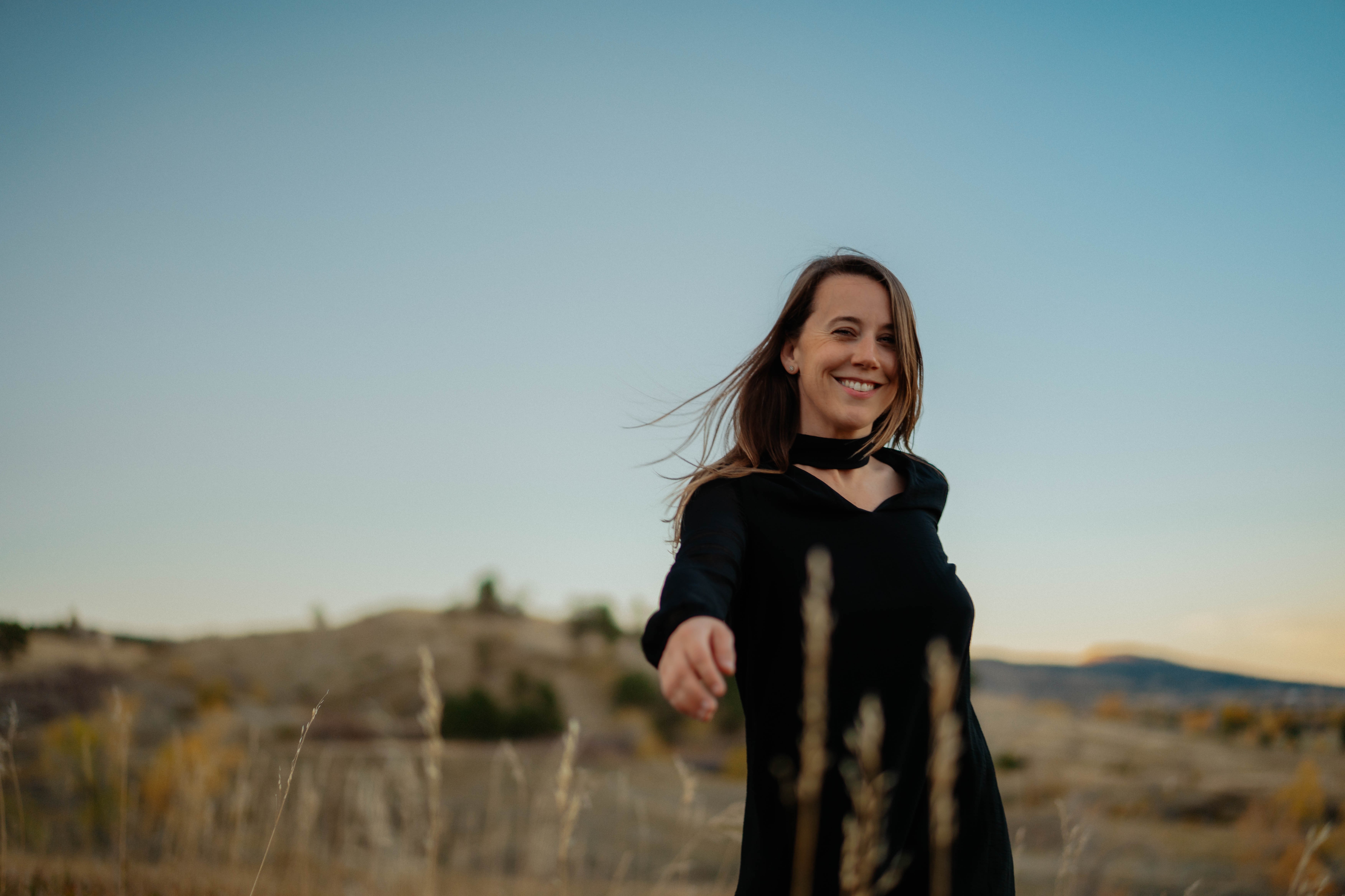 Smiling woman in black dress outdoors with a landscape background, clear sky, and distant hills.