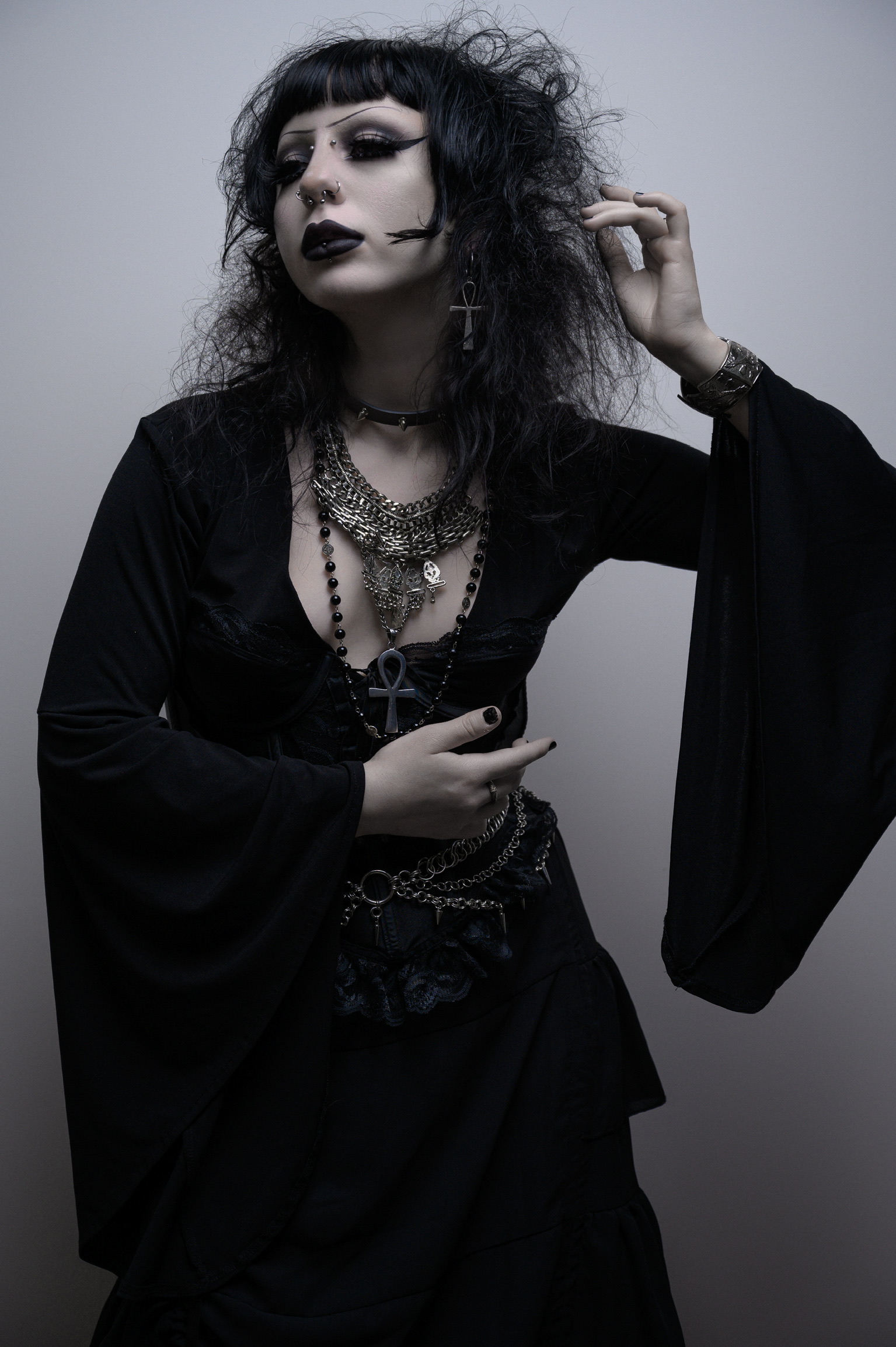Person with dark makeup, curly hair, wearing layered necklaces and black clothing, posing against a plain background.