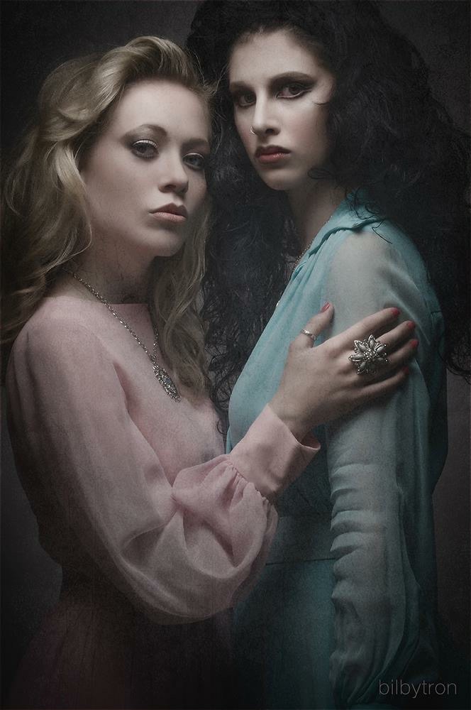 Two women with serious expressions, one with light brown hair in a pink top, the other with dark curly hair in a blue top, close together.