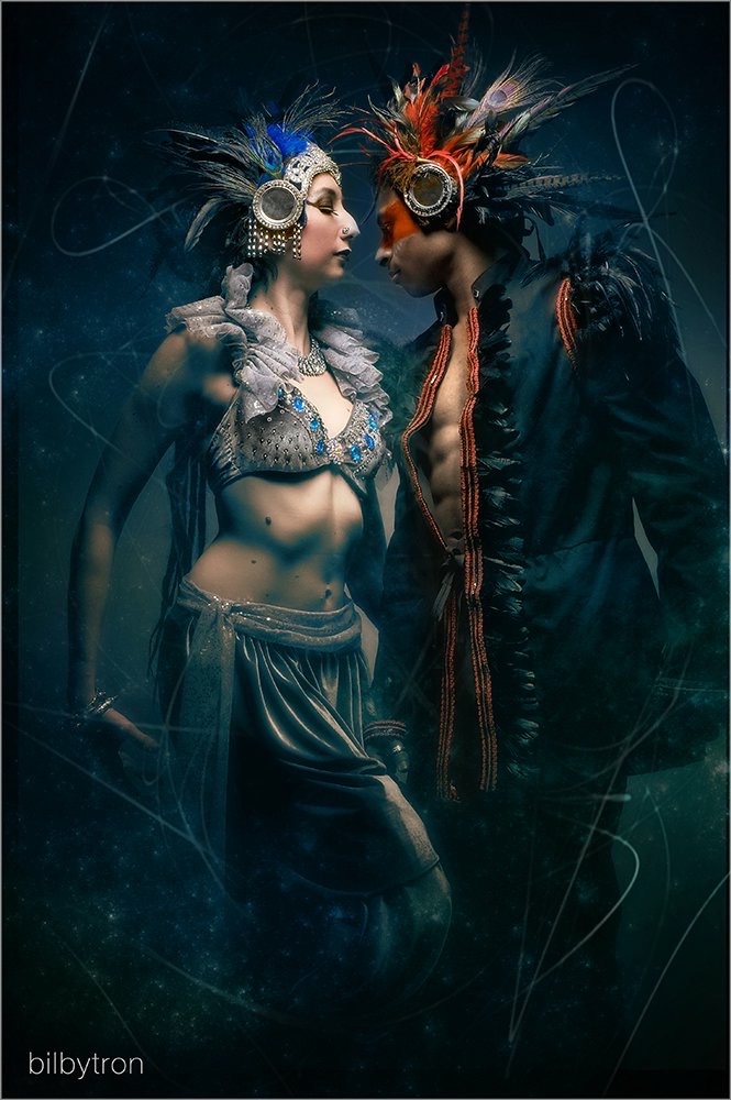 Two performers in elaborate costumes and feathered headdresses face each other against a dark background.