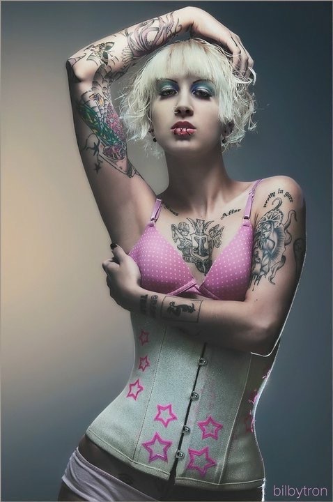 Person with platinum blonde hair, tattoos, and makeup, wearing a pink and white top, posing with arms raised.