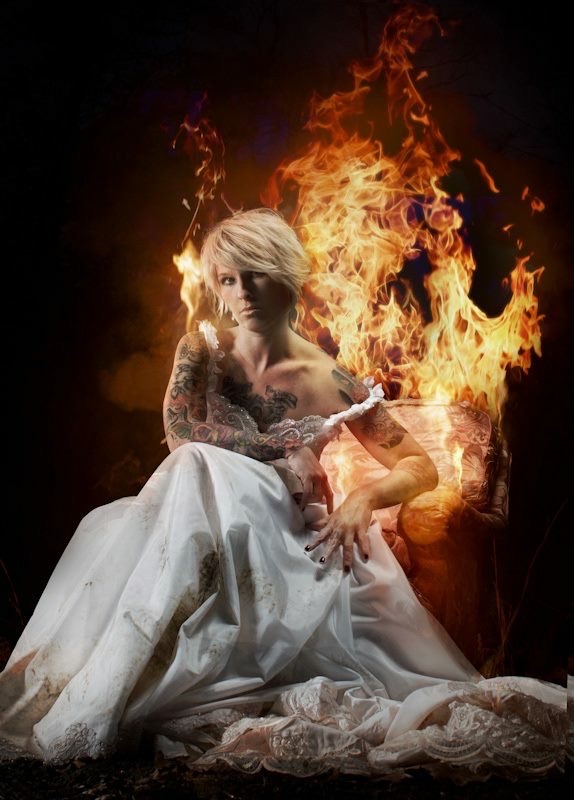 Person with platinum blonde hair in a white dress, surrounded by flames, sitting on the ground with a dark background.