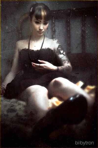 Woman with dark hair and tattoos sitting on a bed, taking a mirror selfie in a dimly lit room.