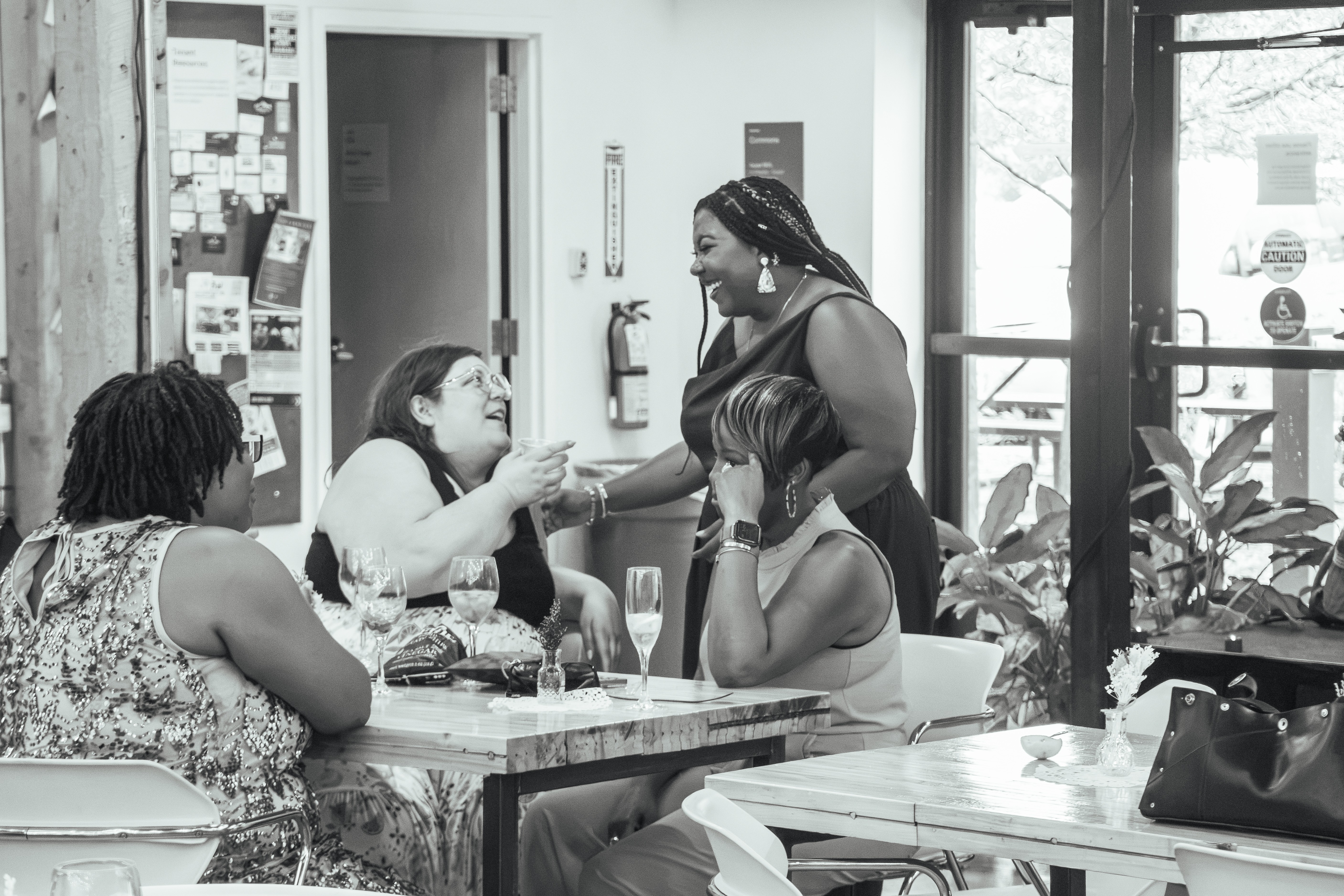 Four women sit at a table in a cafe, with one woman standing and handing something to another woman. Plants and windows are in the background.