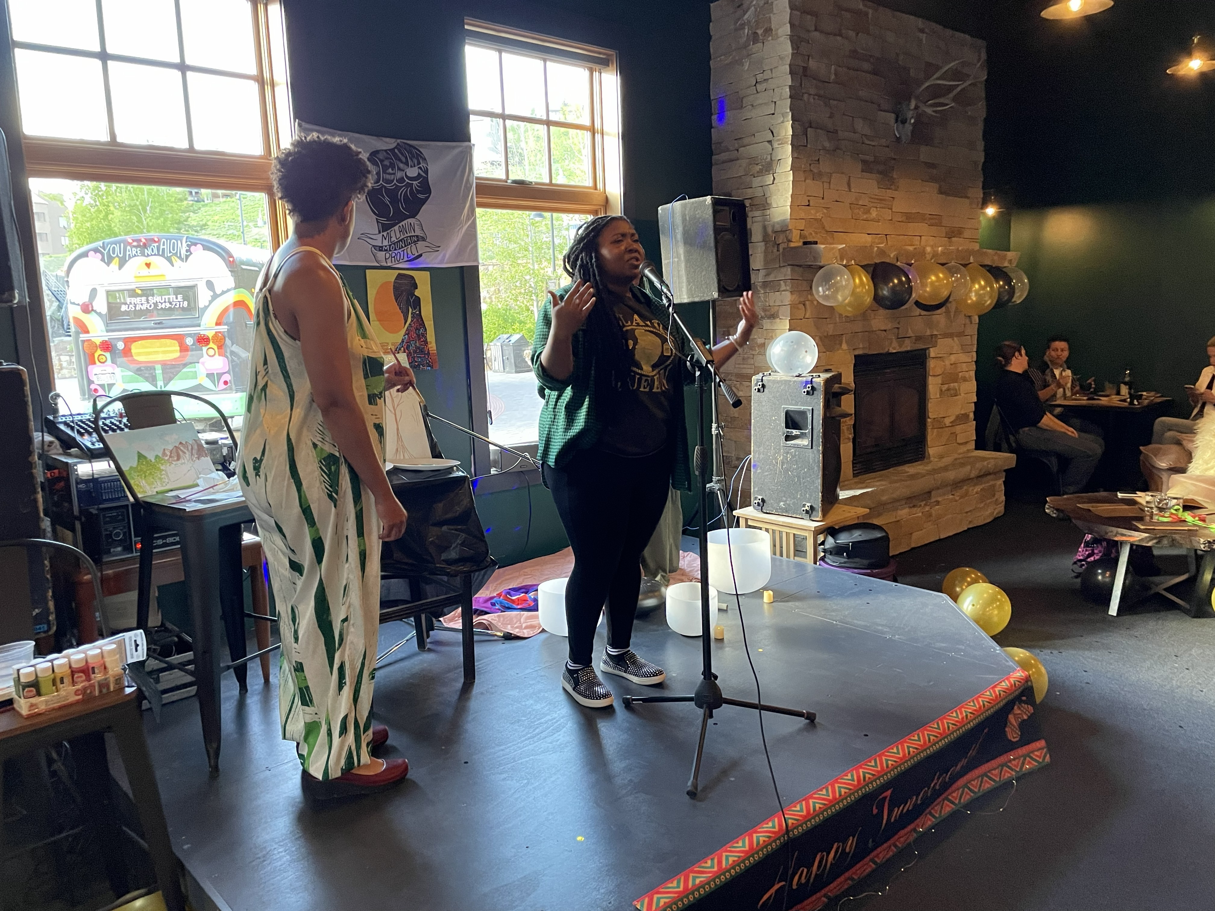 Two women stand on a small stage in a cozy room with large windows, a brick fireplace, and balloons, one speaking into a microphone.