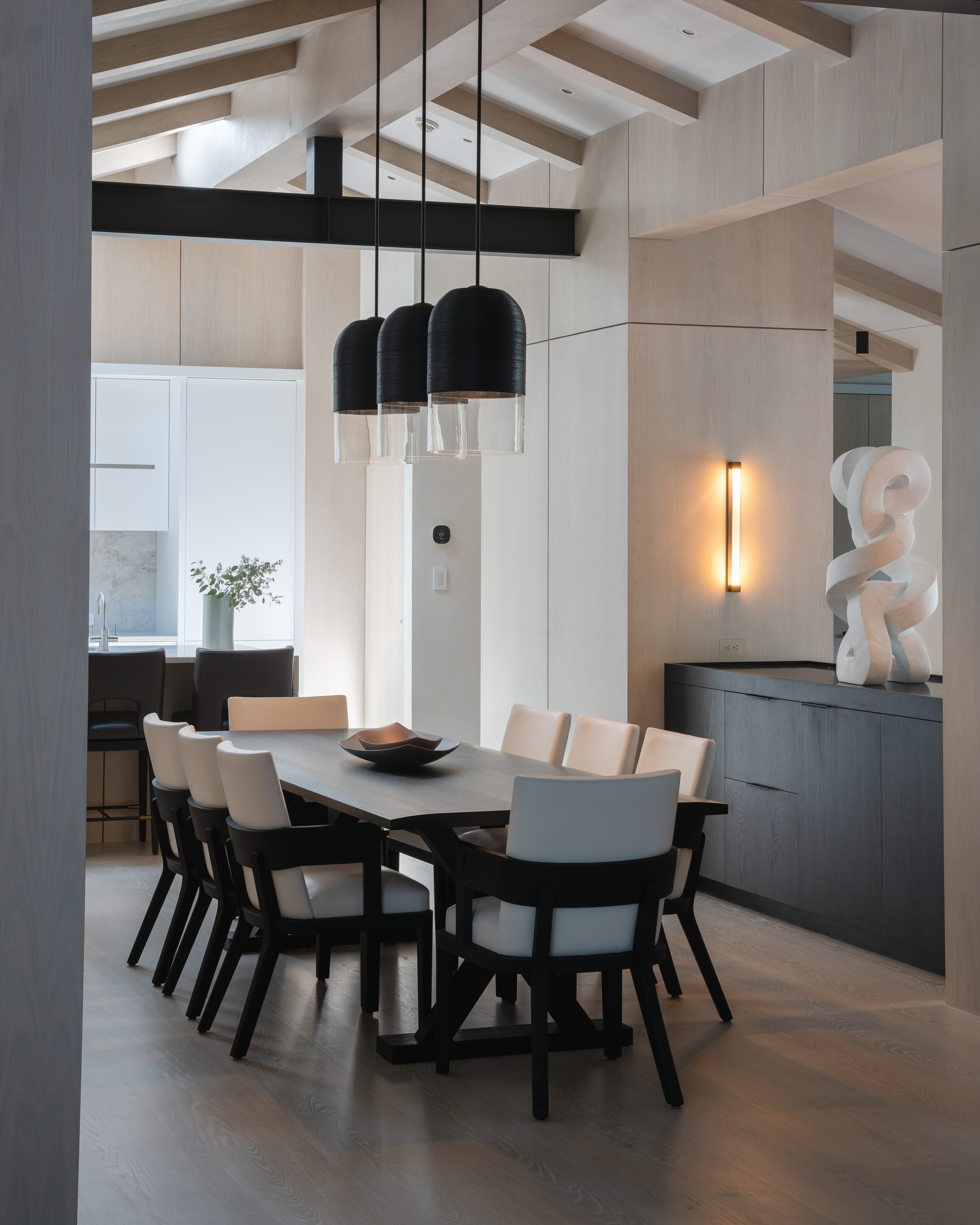 Dining room with a long table, black and white chairs, hanging black pendant lights, and modern decor including a sculpture.