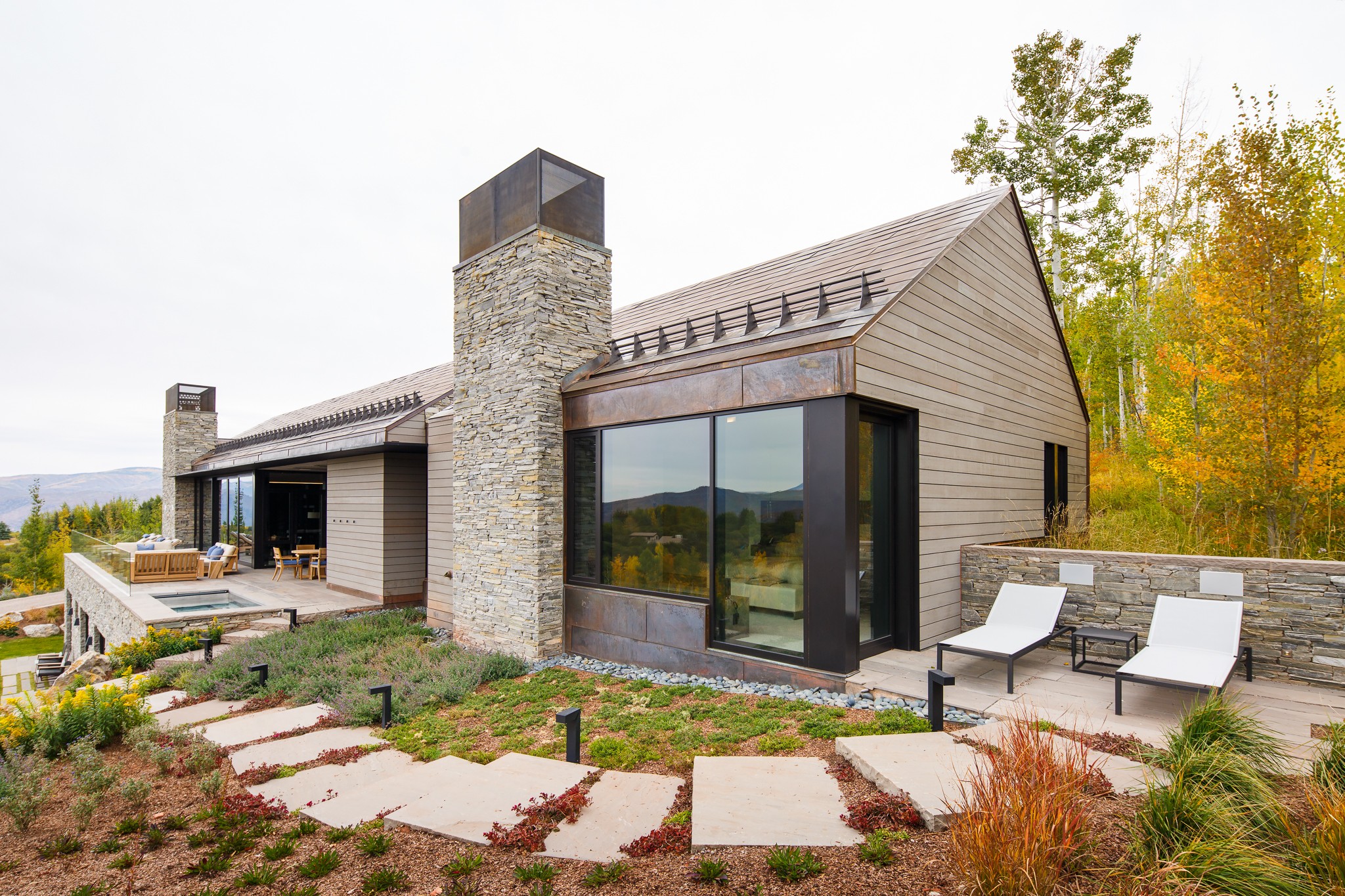 Modern house with stone chimney, large glass windows, outdoor seating, and landscaped garden with trees in autumn colors.