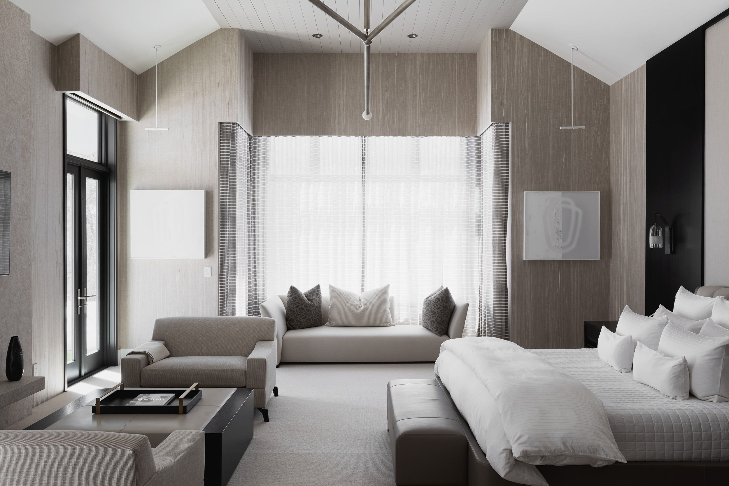 Modern bedroom with large window, white curtains, bed with white linens, and seating area with sofa and pillows.